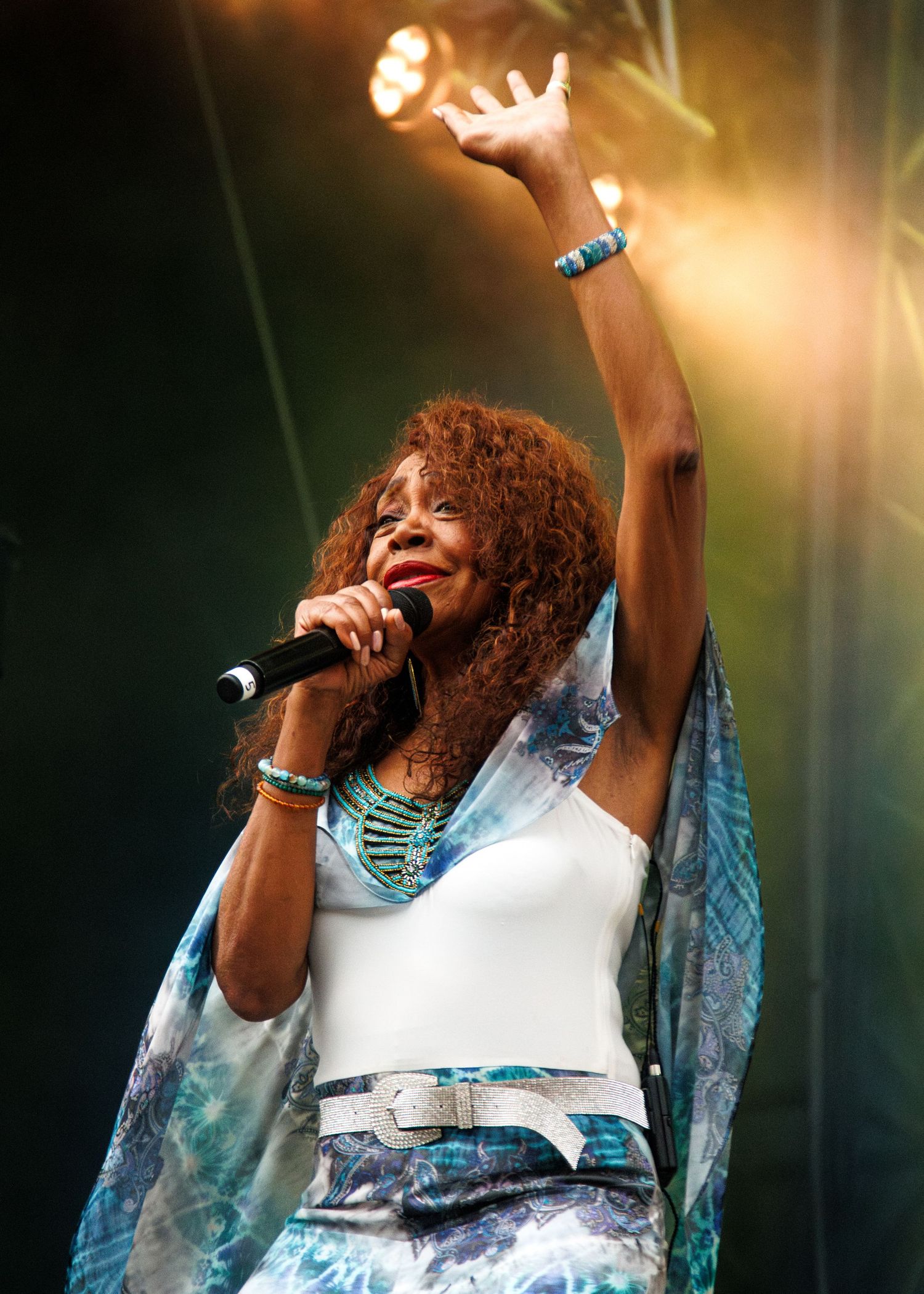 A performer in a white top and blue patterned shawl singing passionately on stage with dramatic lighting effects.