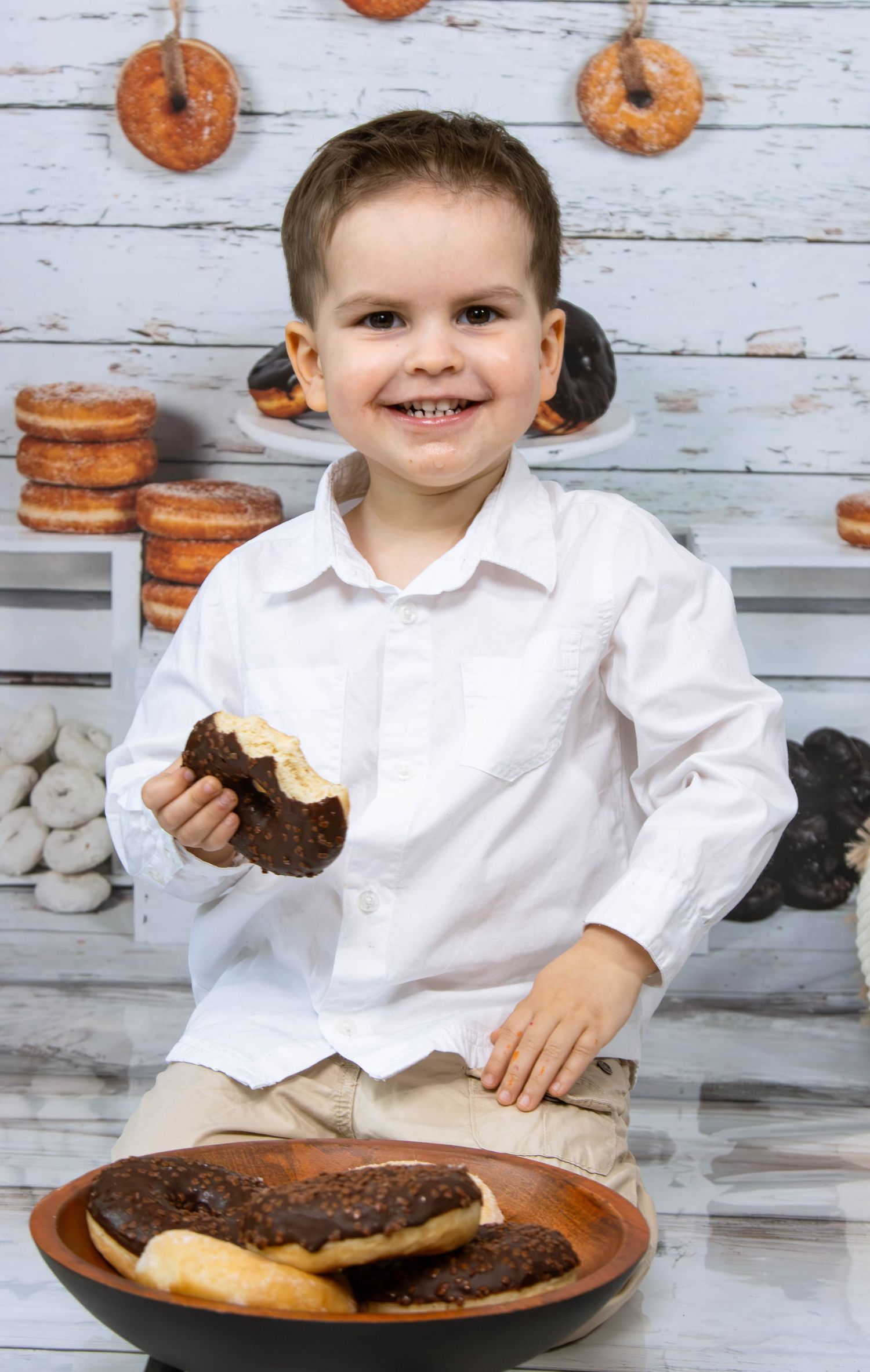 Young child smiling while eating donuts in front of rustic wood backdrop with decorative pastries.