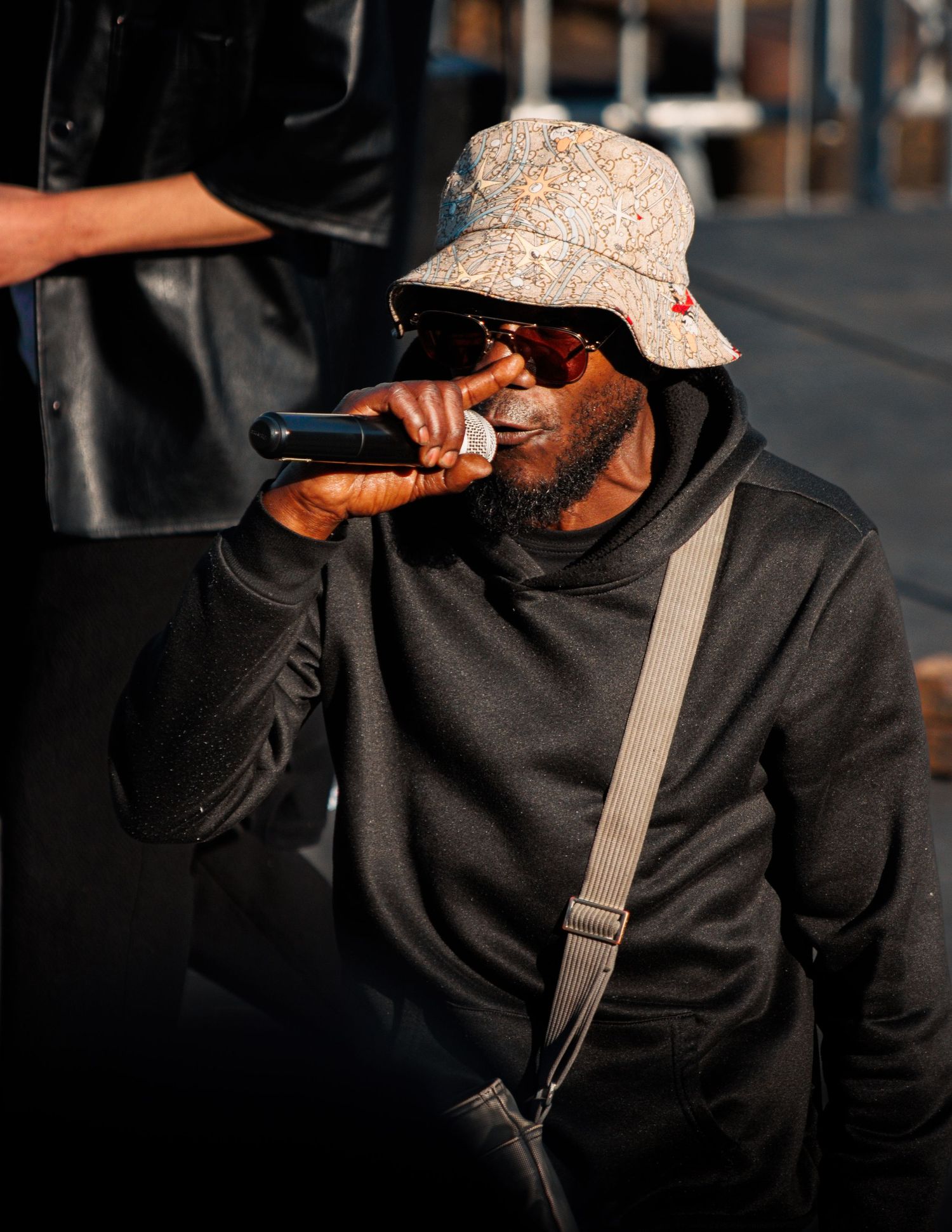 Performer in bucket hat and sunglasses singing into microphone outdoors.