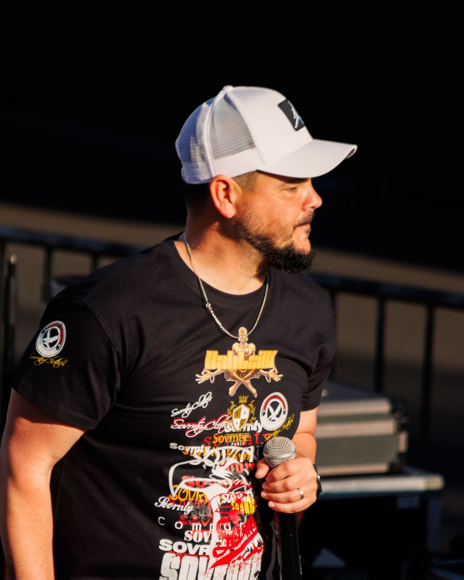 Performer wearing baseball cap and graphic t-shirt holds microphone during outdoor show.