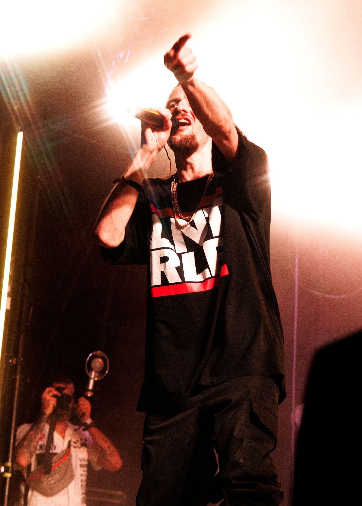 Energetic performer in black clothing sings passionately on stage with dramatic lighting effects in the background.