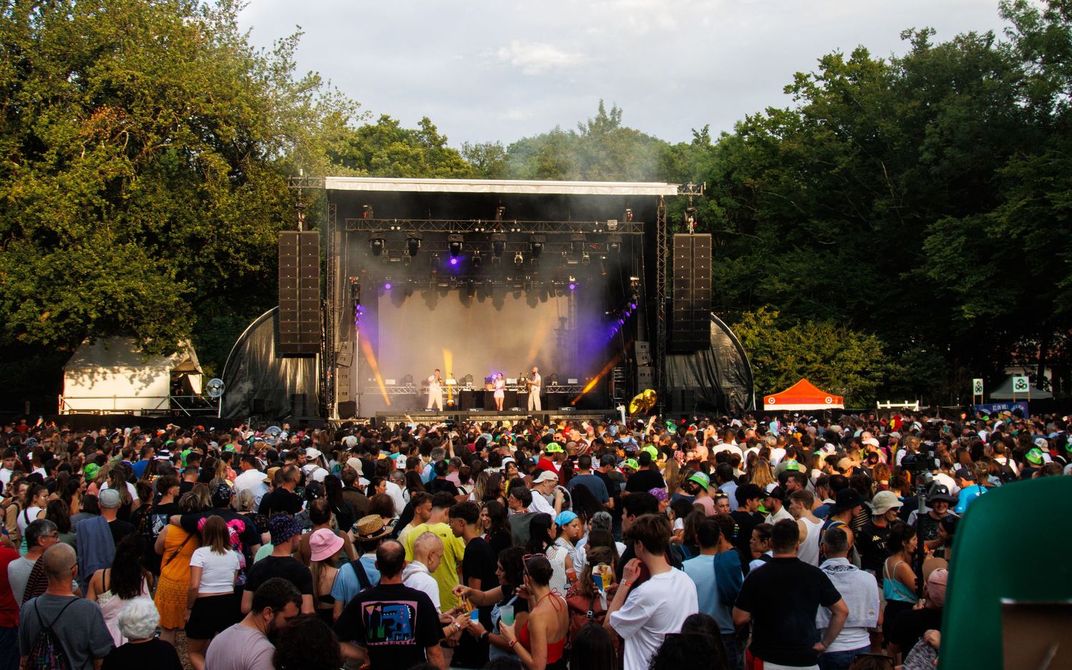 A large crowd gathers at an outdoor music festival with stage lights and trees visible in the background.