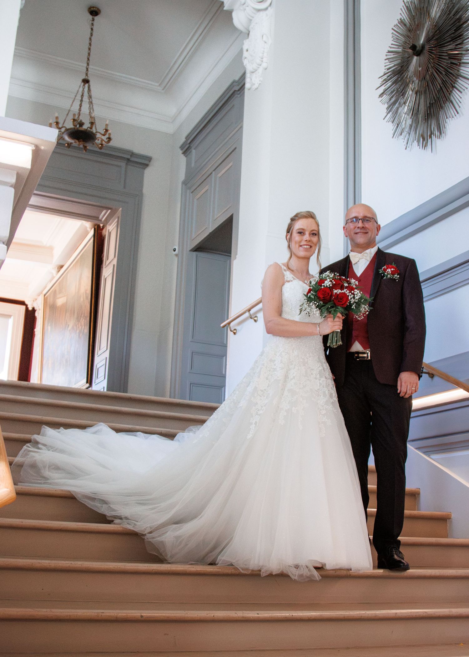 Bride in lace wedding gown with long train stands with groom on elegant staircase holding red bouquet.