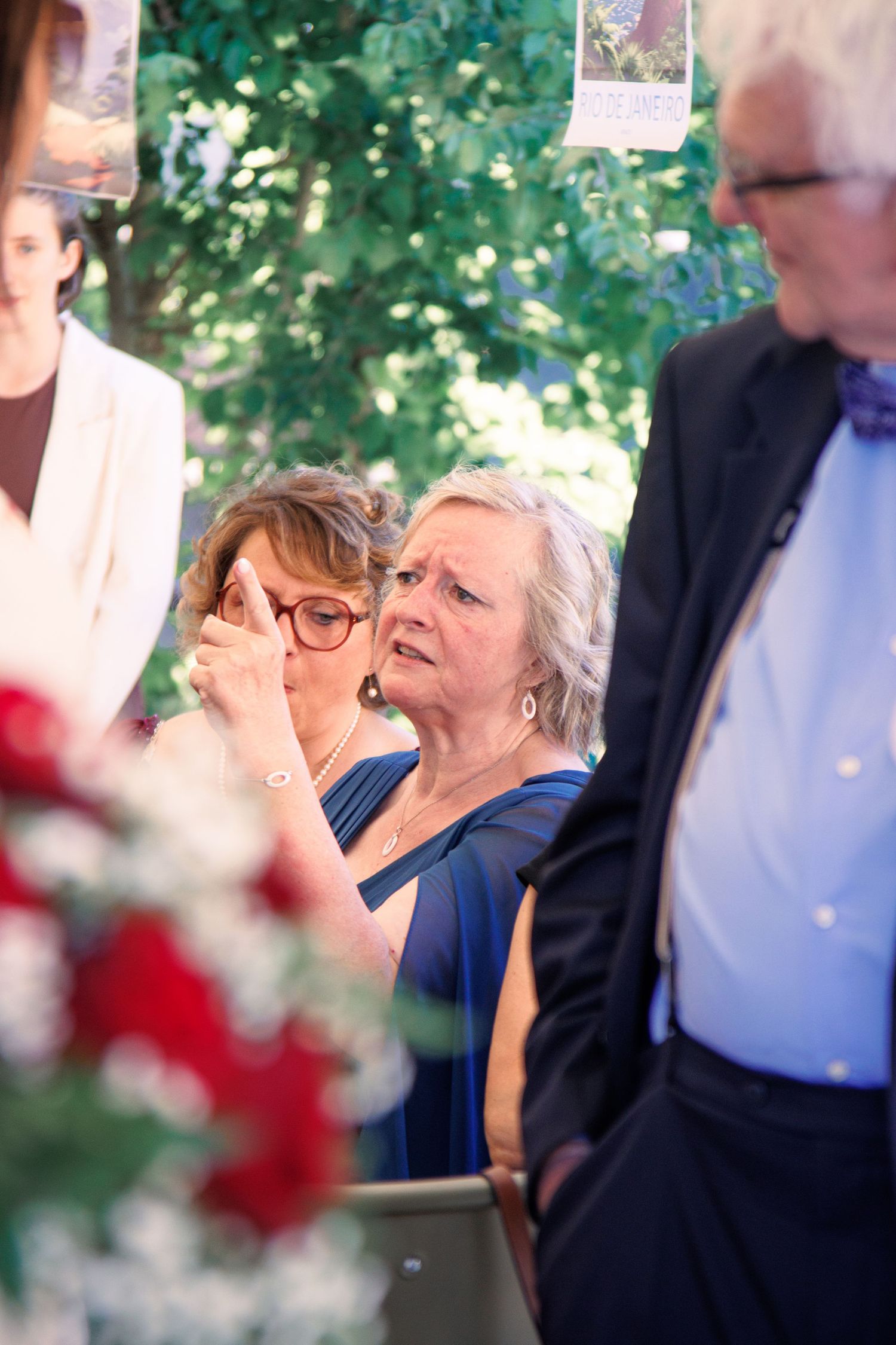 Two emotional female wedding guests wiping tears during outdoor wedding ceremony surrounded by red floral arrangements.