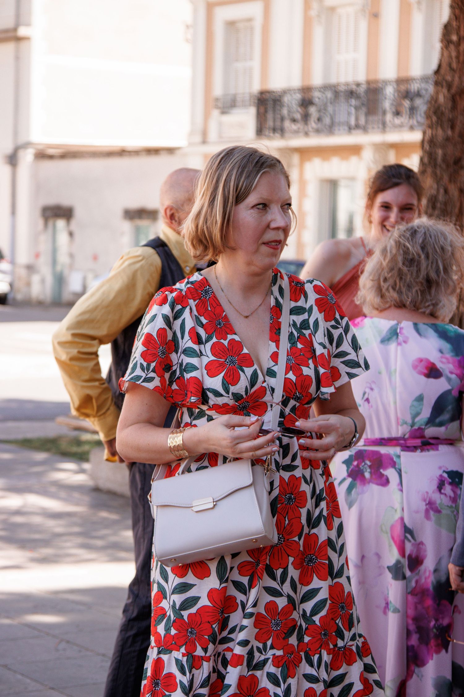 Woman in red floral dress with white handbag socializing outdoors at a sunny event.