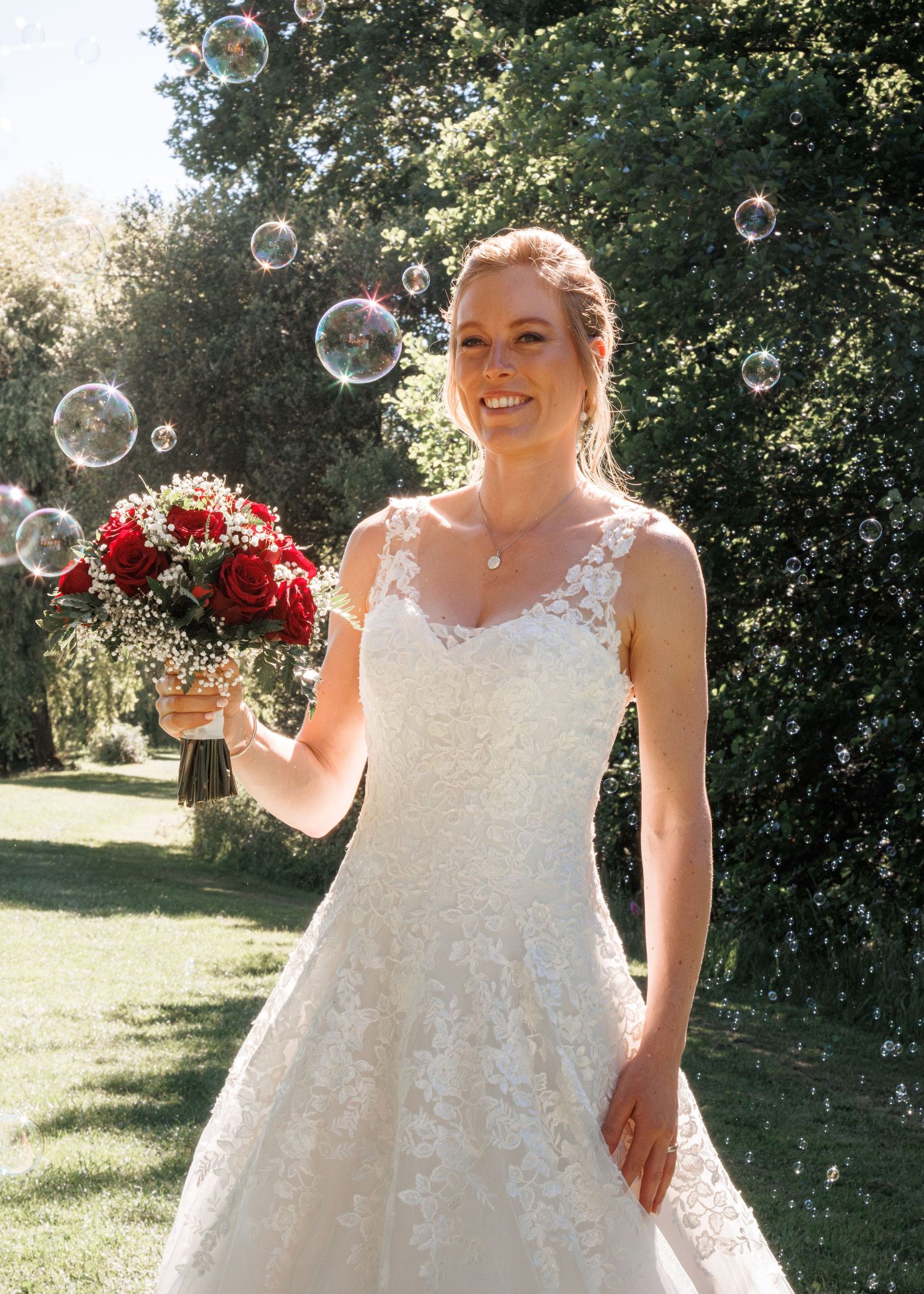 Smiling bride in lace gown holding red rose bouquet surrounded by floating bubbles outdoors.