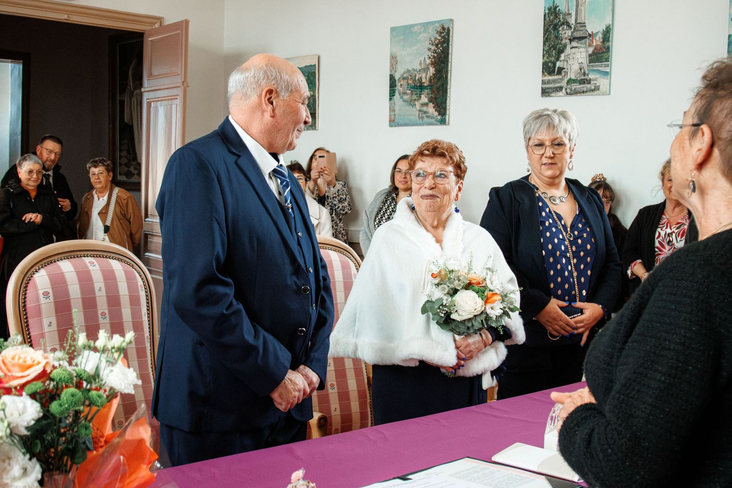 Elderly couple exchanging vows at an intimate wedding ceremony, bride holding a bouquet with guests watching.