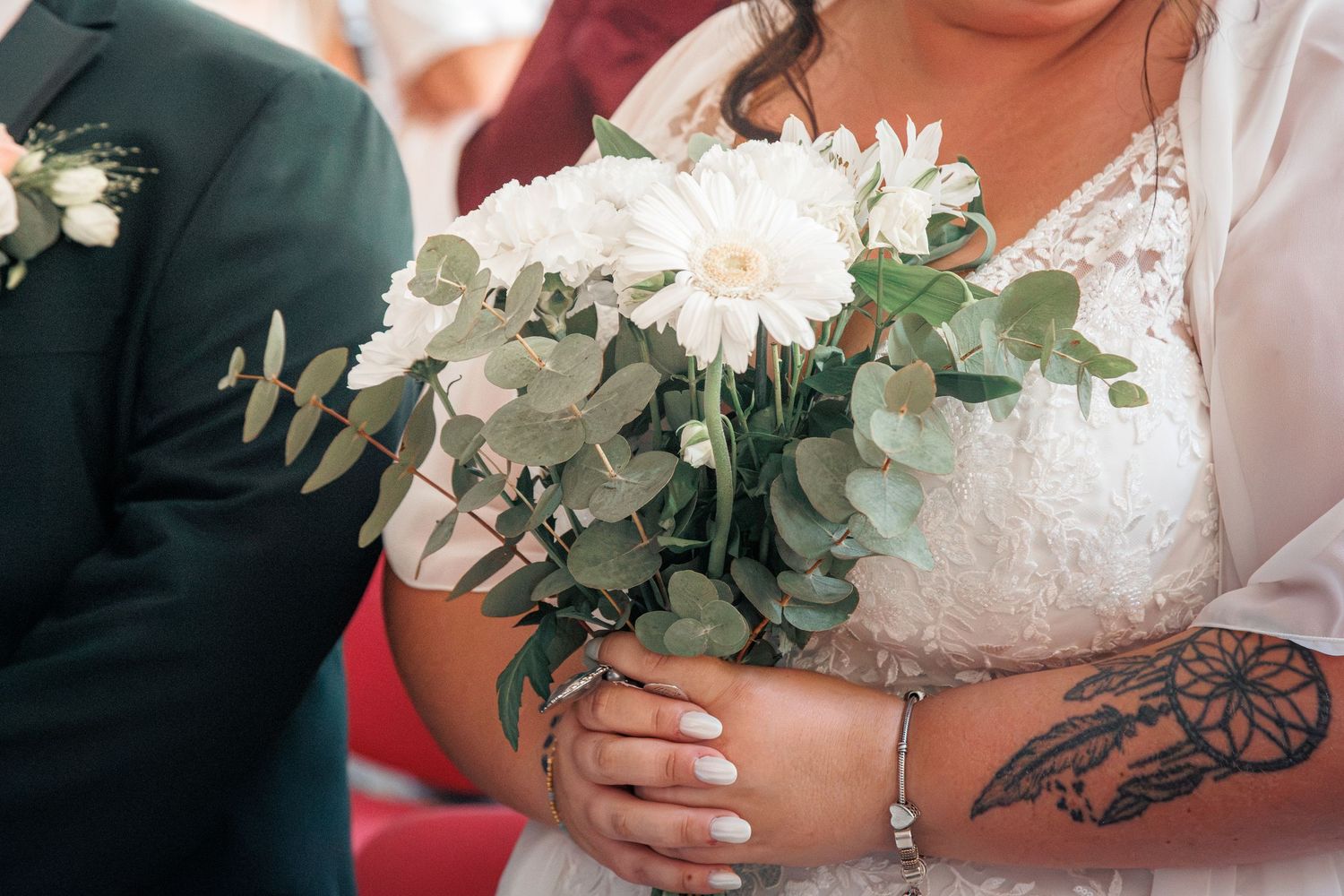 Bride holding white gerbera daisy and eucalyptus bouquet with tattoo visible on wrist.