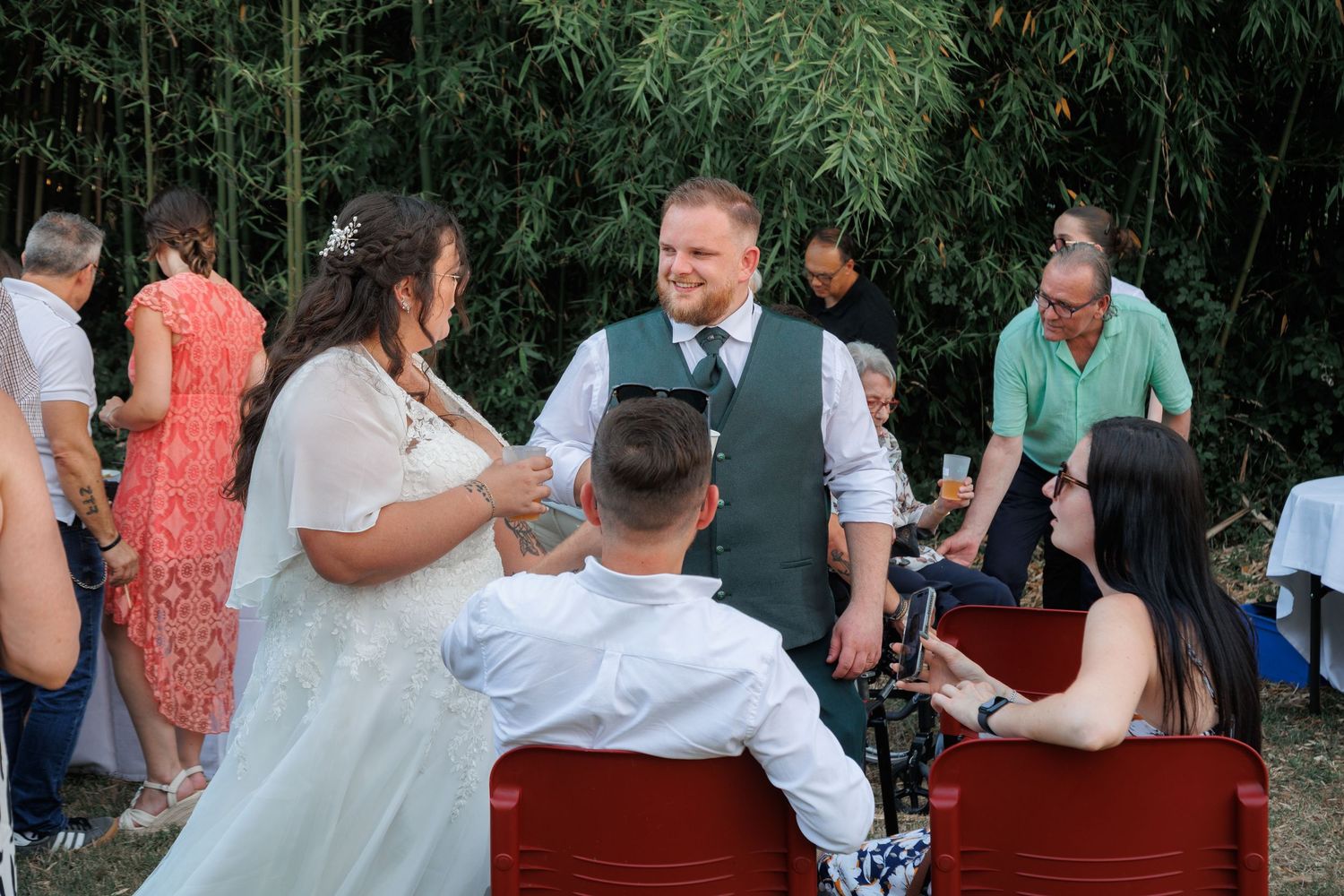 Bride and groom in green vest chat with seated guests at outdoor wedding.