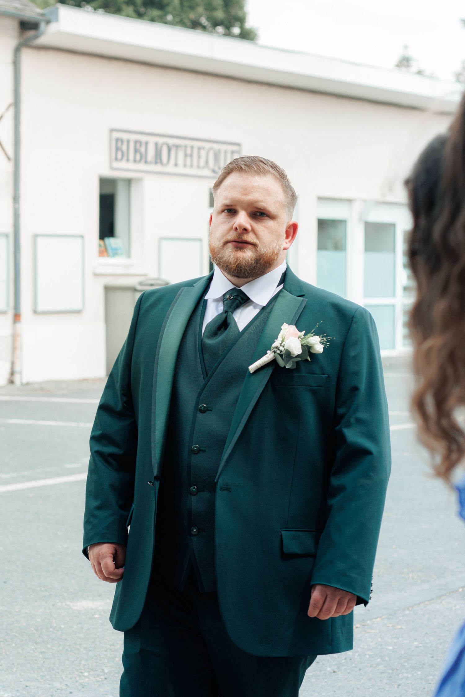 Groom in dark green three-piece suit with boutonniere stands outside a bibliotheque building.