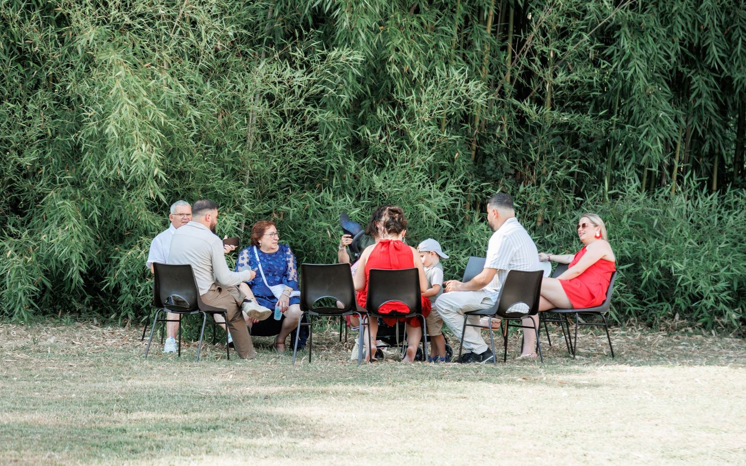 Group of wedding guests seated in circle of chairs outdoors near tall green bamboo trees.
