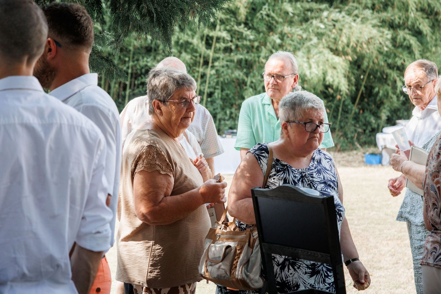 Wedding guests mingling outdoors in summer, two older women conversing at garden cocktail reception.