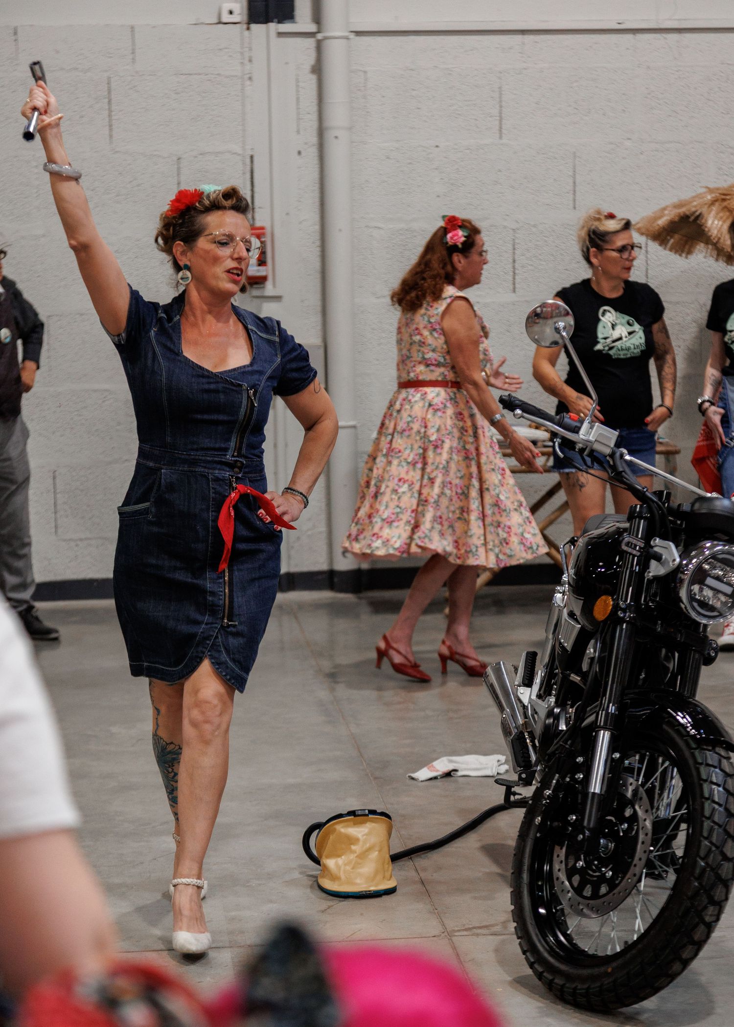 Women in vintage 1950s pin-up style dresses pose near a motorcycle at an indoor retro-themed event.