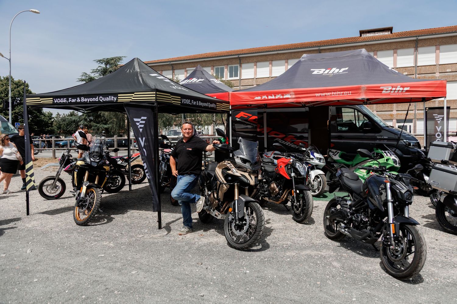 Motorcycle exhibition with colorful bikes displayed under branded tents at an outdoor event in a sunny parking area.