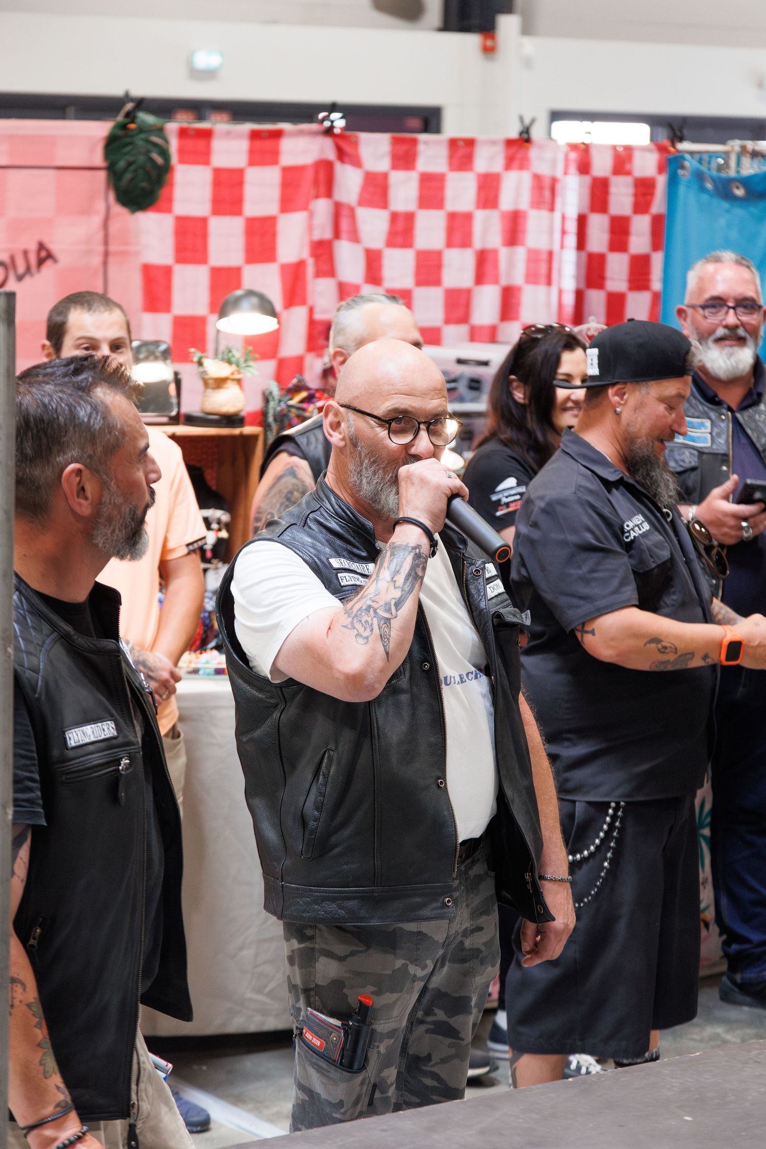 Tattooed biker in leather vest speaks into microphone at an indoor motorcycle club gathering with members around him.