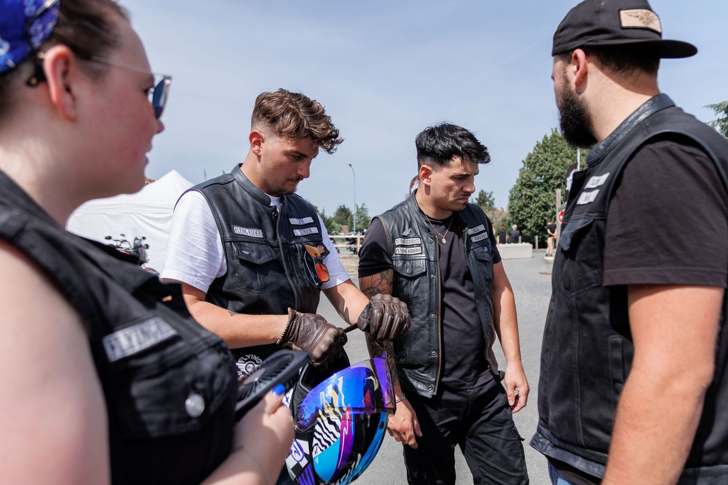 Motorcycle club members in vests signing a helmet outdoors on a sunny day.
