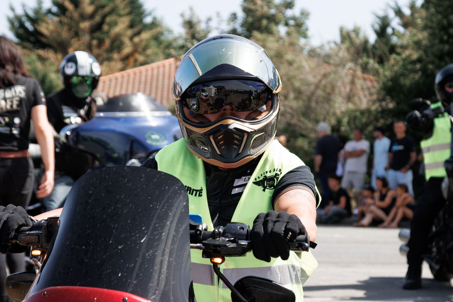 Motorcyclist wearing a green vest, full-face helmet, and gloves prepares to ride at an outdoor motorsport event.