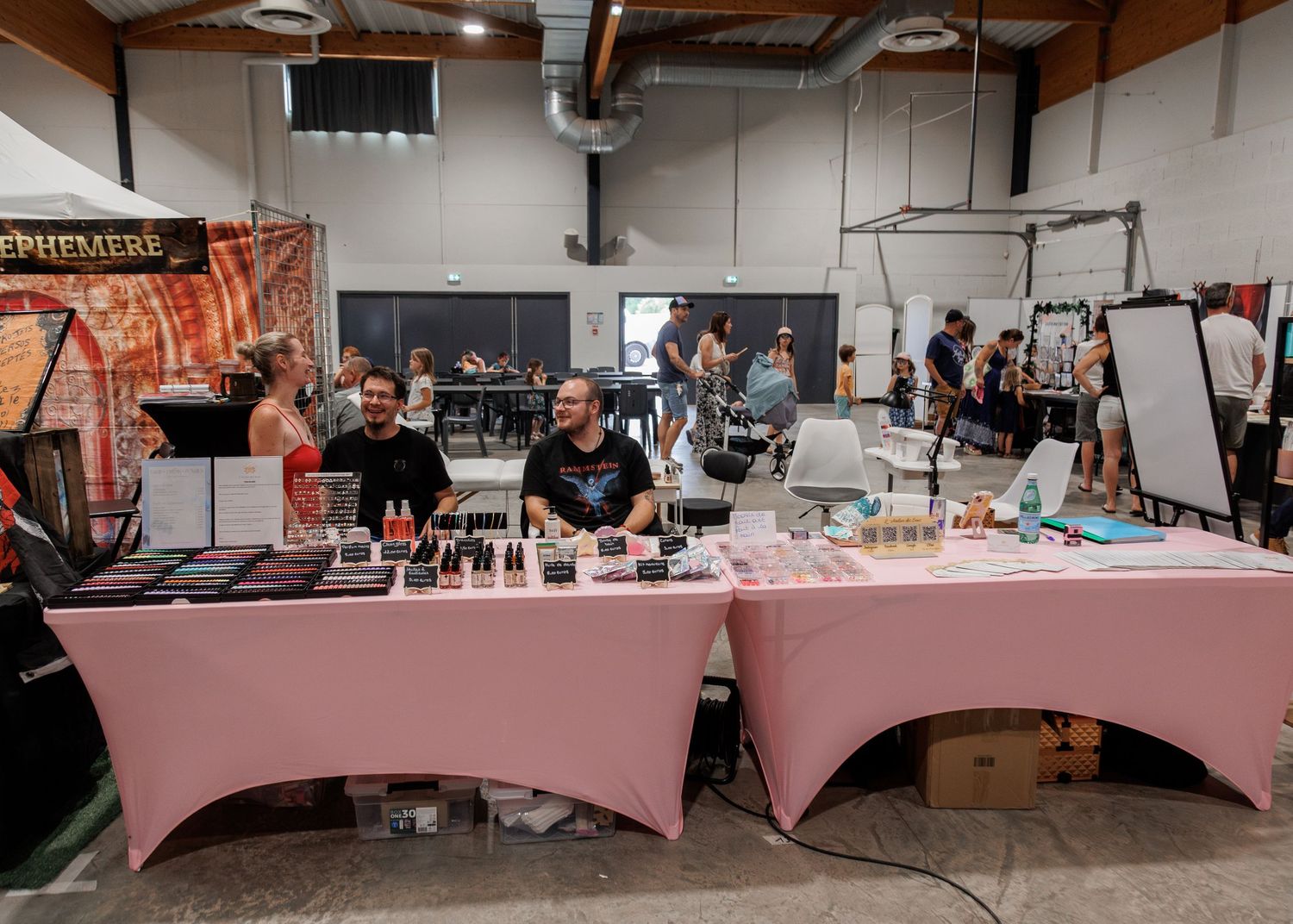 Vendors smile behind a pink draped table displaying makeup products at an indoor trade show event.