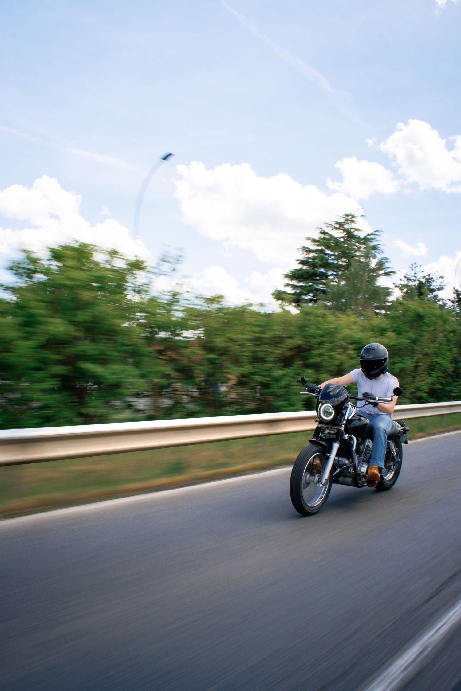 Motorcyclist riding a classic motorcycle at speed on a highway, wearing helmet and jeans, with blurred green trees and blue sky.