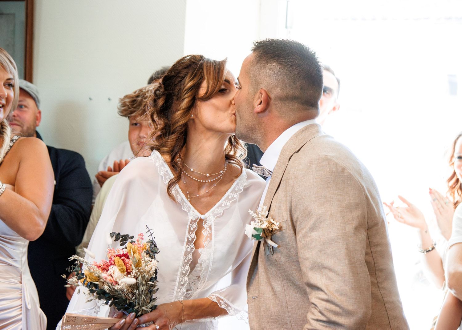 Newlyweds share a romantic kiss while guests celebrate with joyful expressions during a wedding reception.