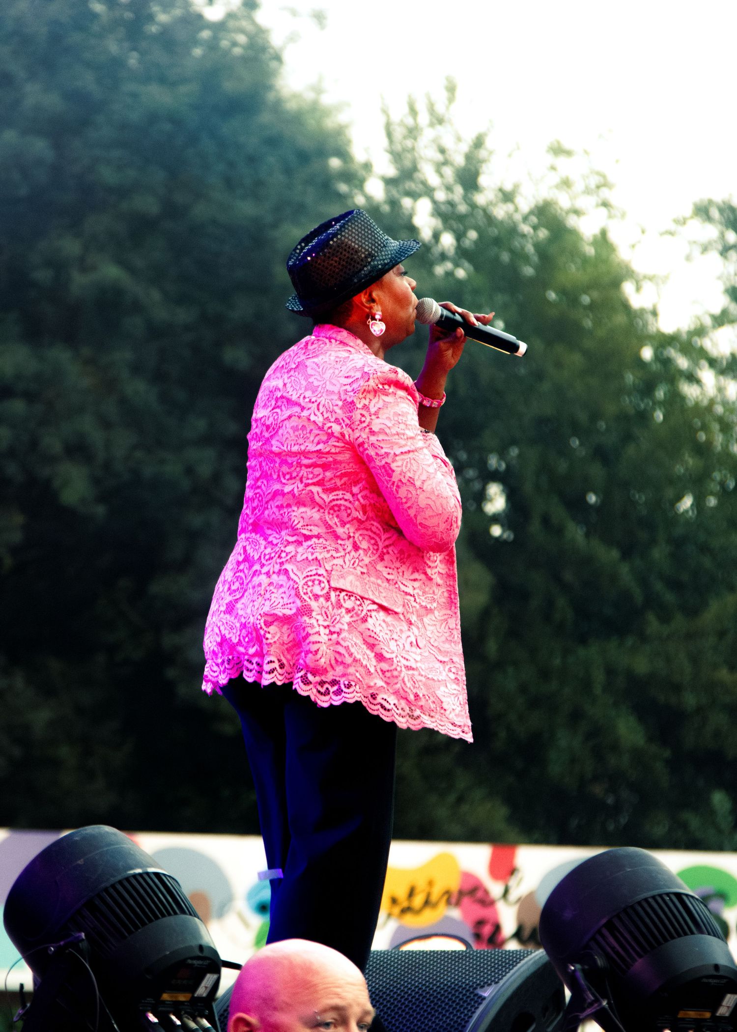 A performer in a bright pink lace top and black hat sings on stage while holding a microphone during an outdoor concert.