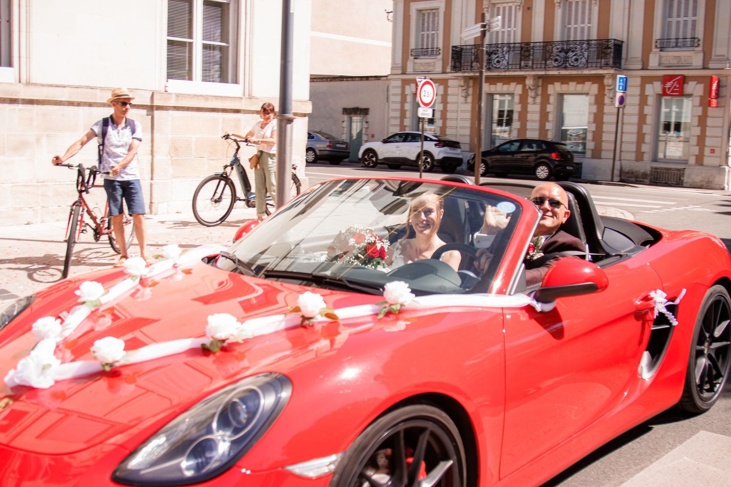 Newlyweds celebrating in decorated red Porsche convertible outside French wedding venue.