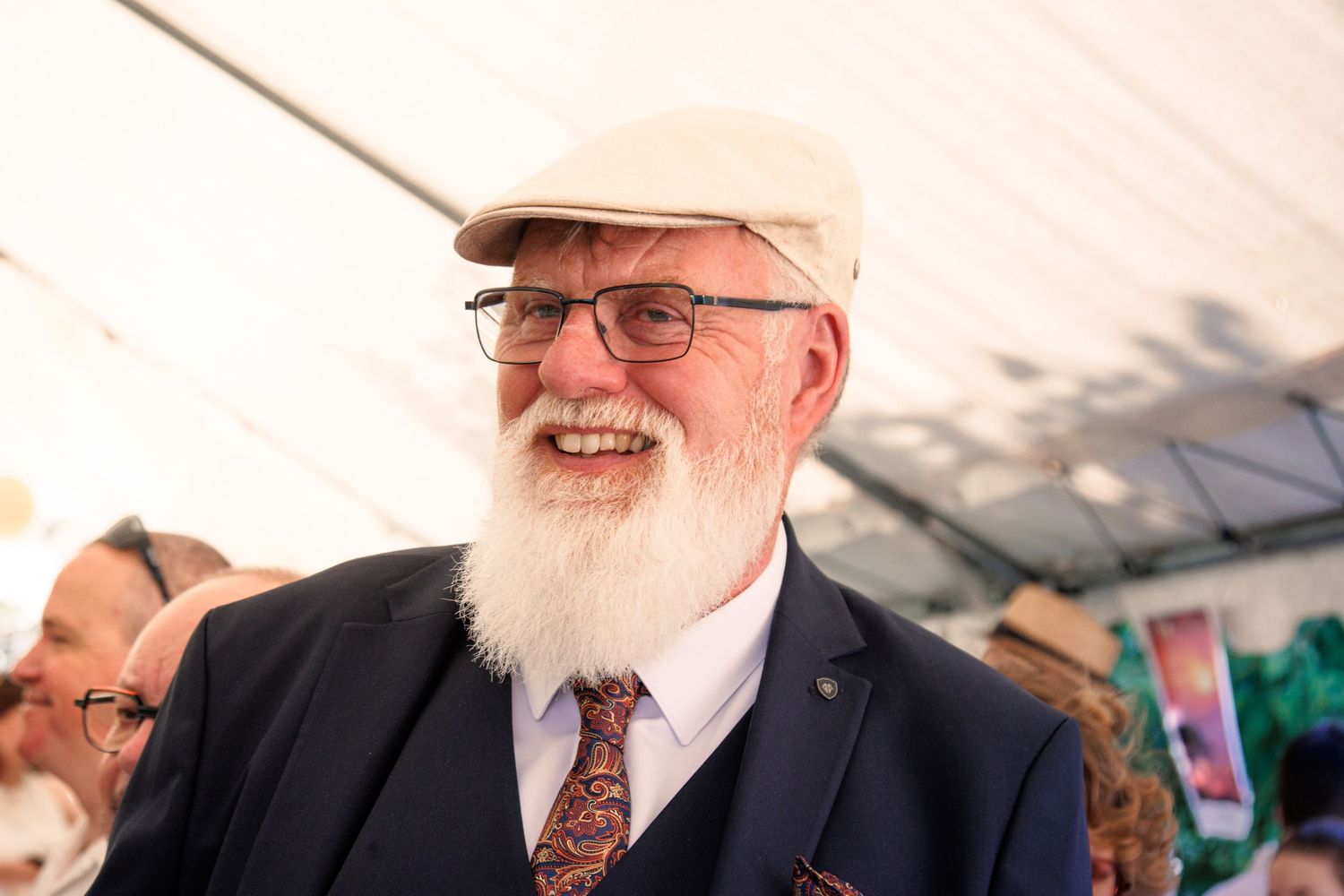 Cheerful elderly man with white beard and flat cap smiles at outdoor wedding reception under tent.