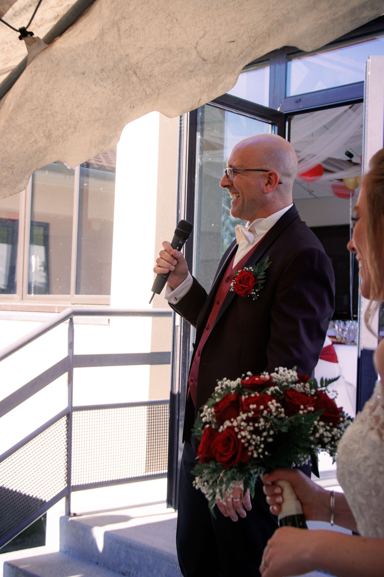 Bald groom in dark suit with red boutonniere speaking into microphone at wedding ceremony holding red rose bouquet.