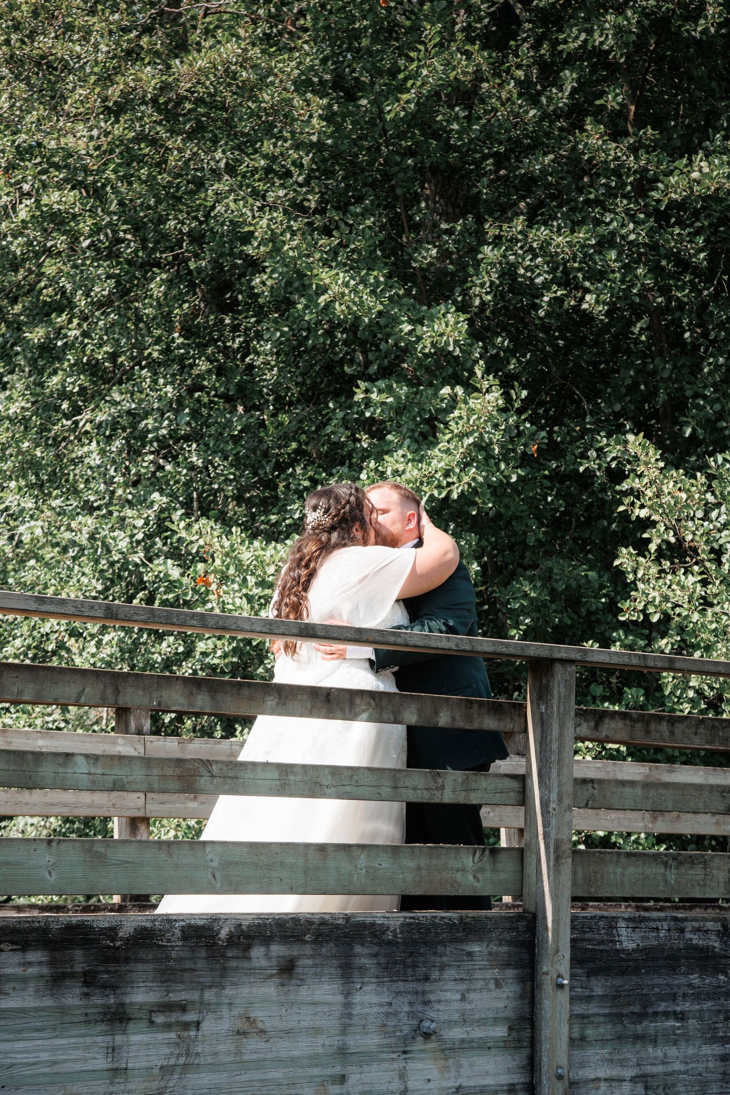 A bride and groom share a romantic embrace on a wooden bridge surrounded by lush green trees on their wedding day.