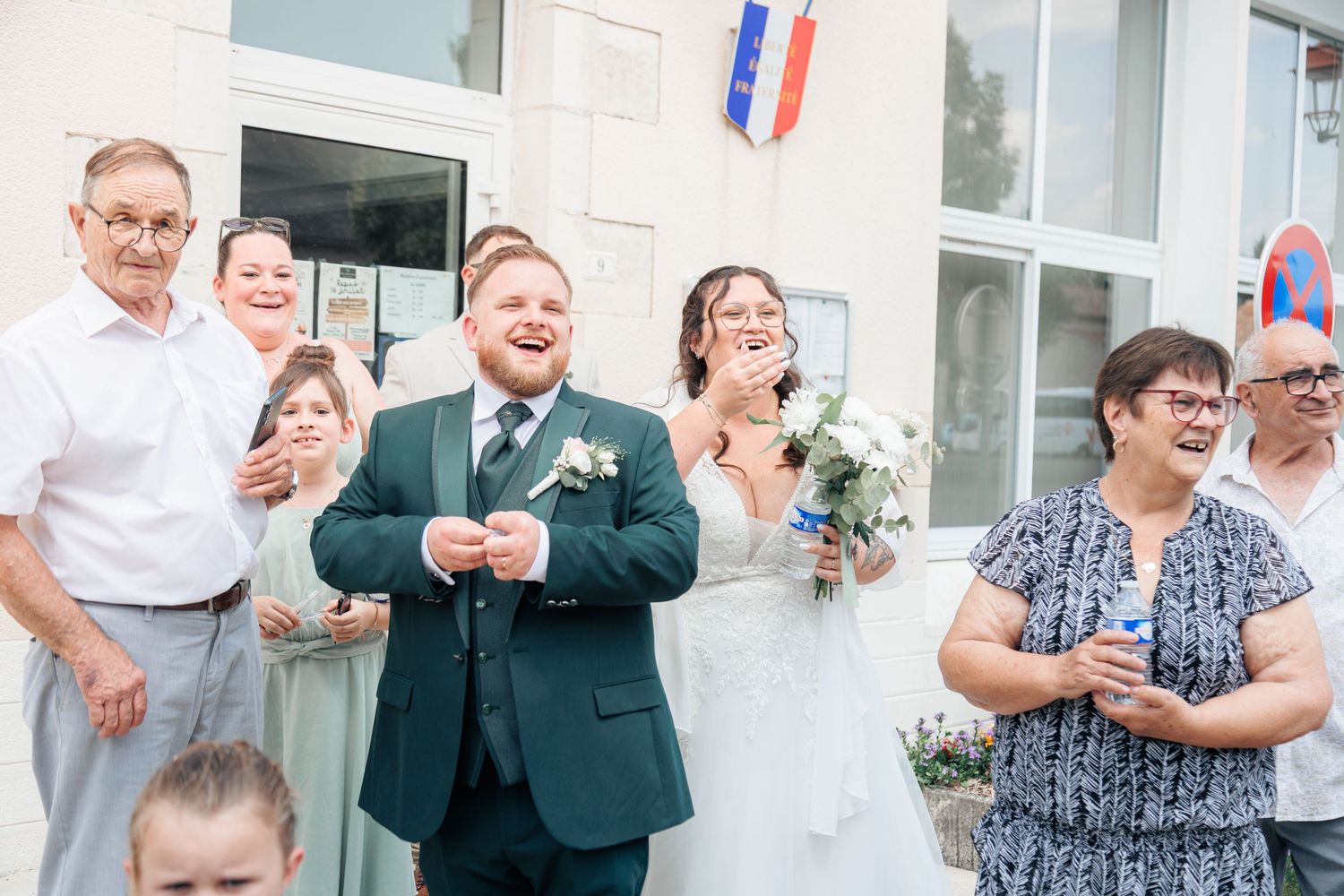 Joyful wedding couple laughing outside a building with French flag, surrounded by celebrating guests.