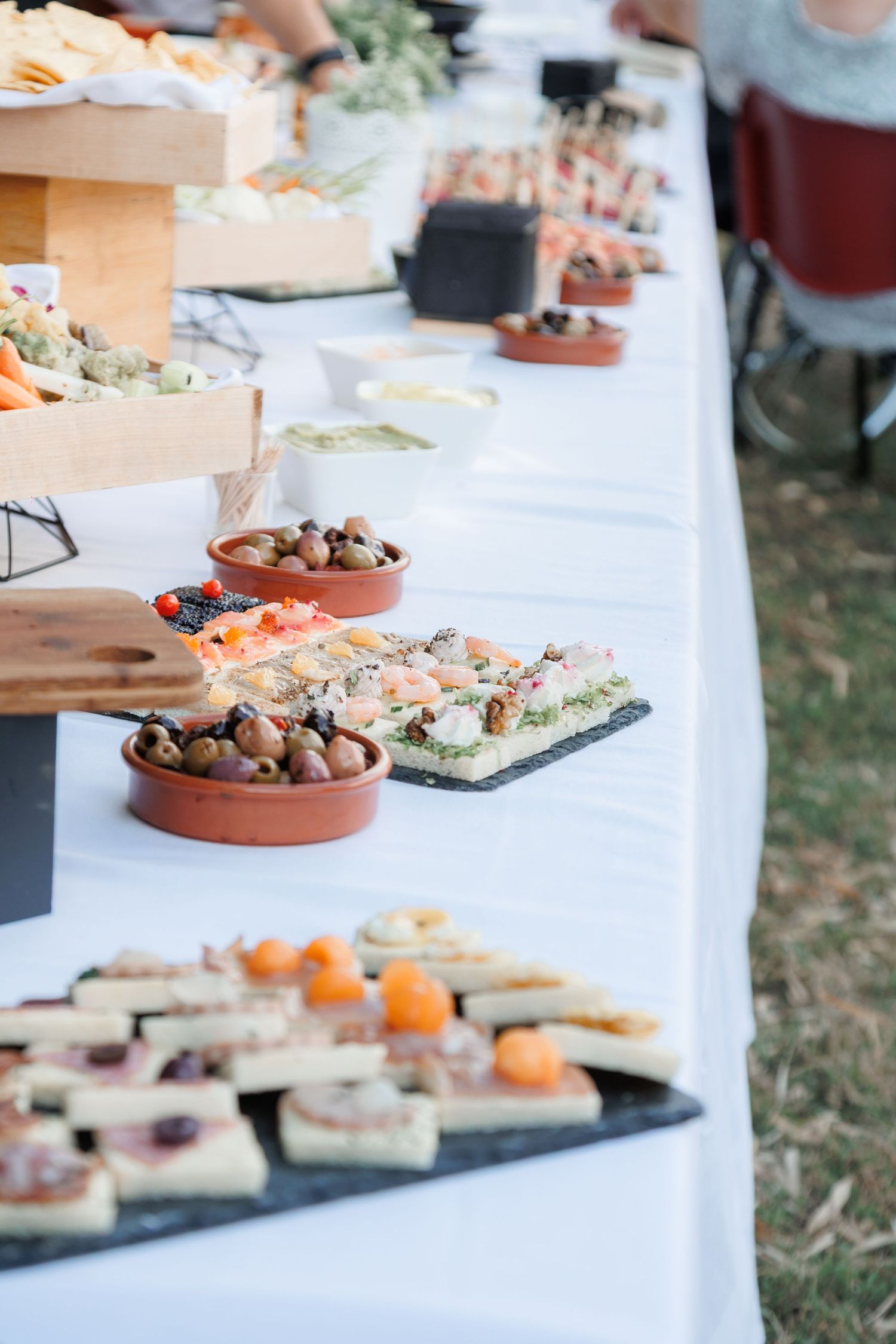 Elegant outdoor wedding reception buffet table with assorted appetizers, olives, and colorful canapes on white linen.