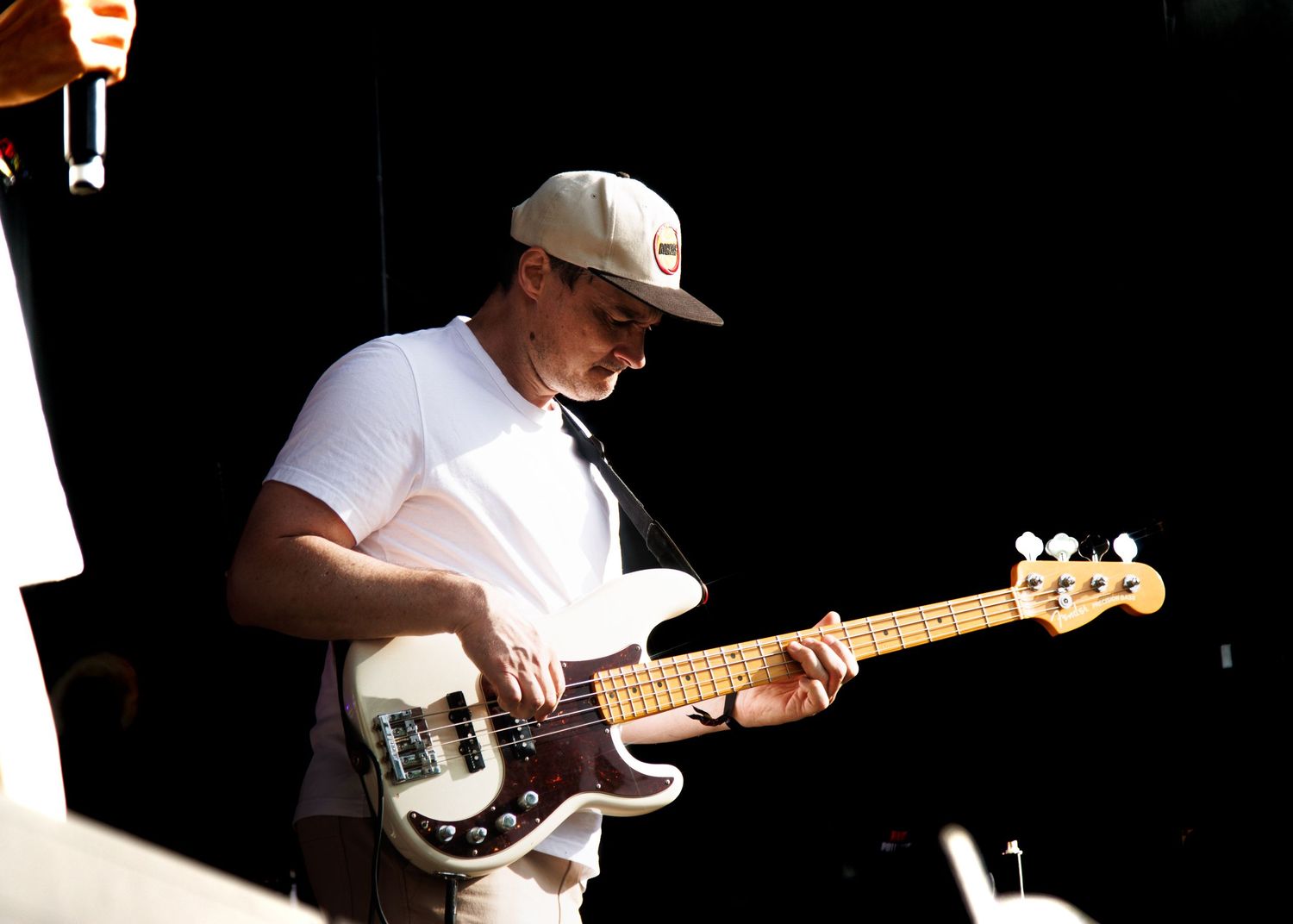 Bass guitarist performing on stage in a white t-shirt against a dark background with dramatic lighting.