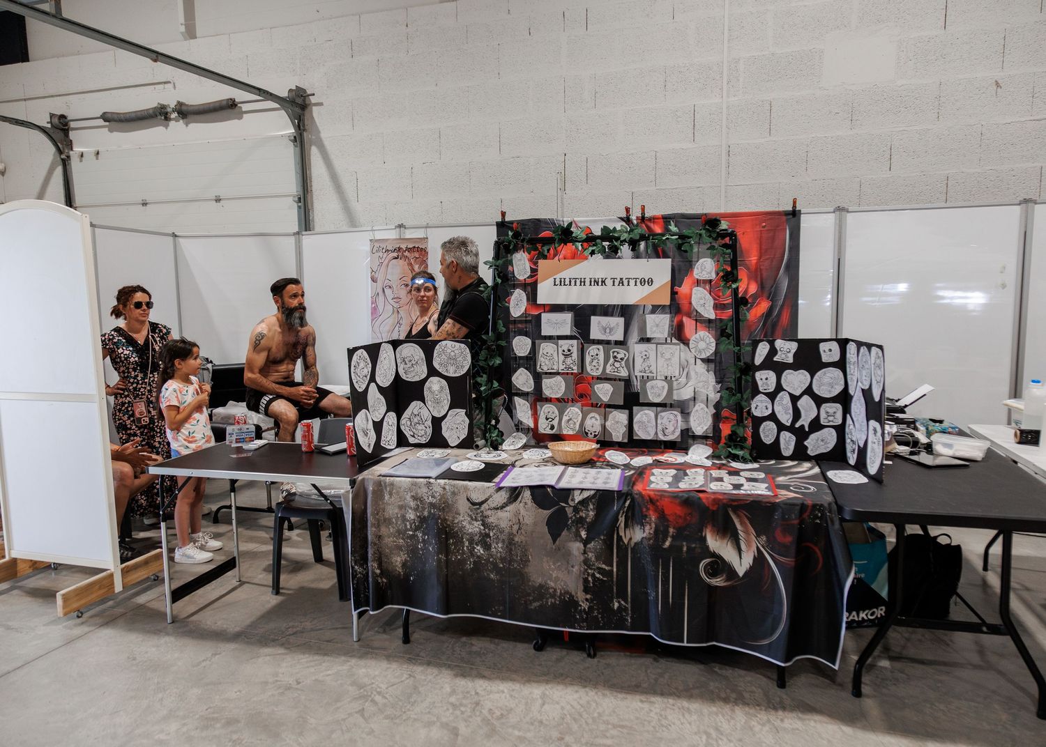 Tattoo convention booth display with flash art sheets, dark tablecloth, and artists consulting with clients indoors.