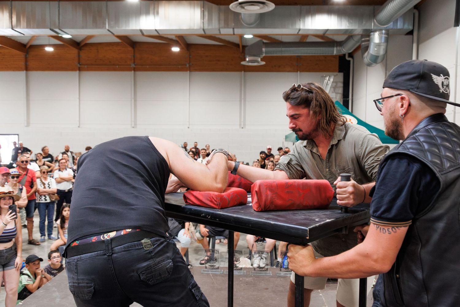 Two men compete in an arm wrestling match on a padded table at an indoor event with a crowd watching.