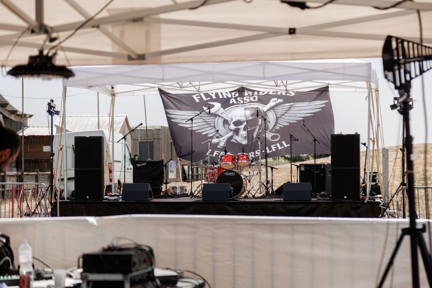 Outdoor stage setup with Flying Riders Association banner, drum kit, and speakers under a white tent canopy.