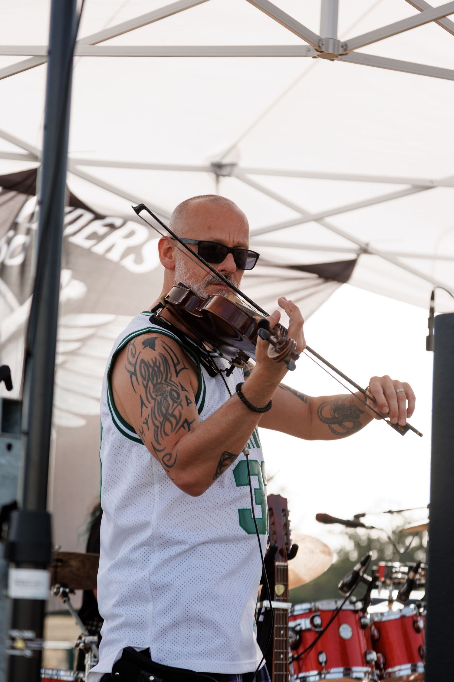 Tattooed bald man playing violin outdoors at a live music event, wearing a white jersey and sunglasses under a white tent.