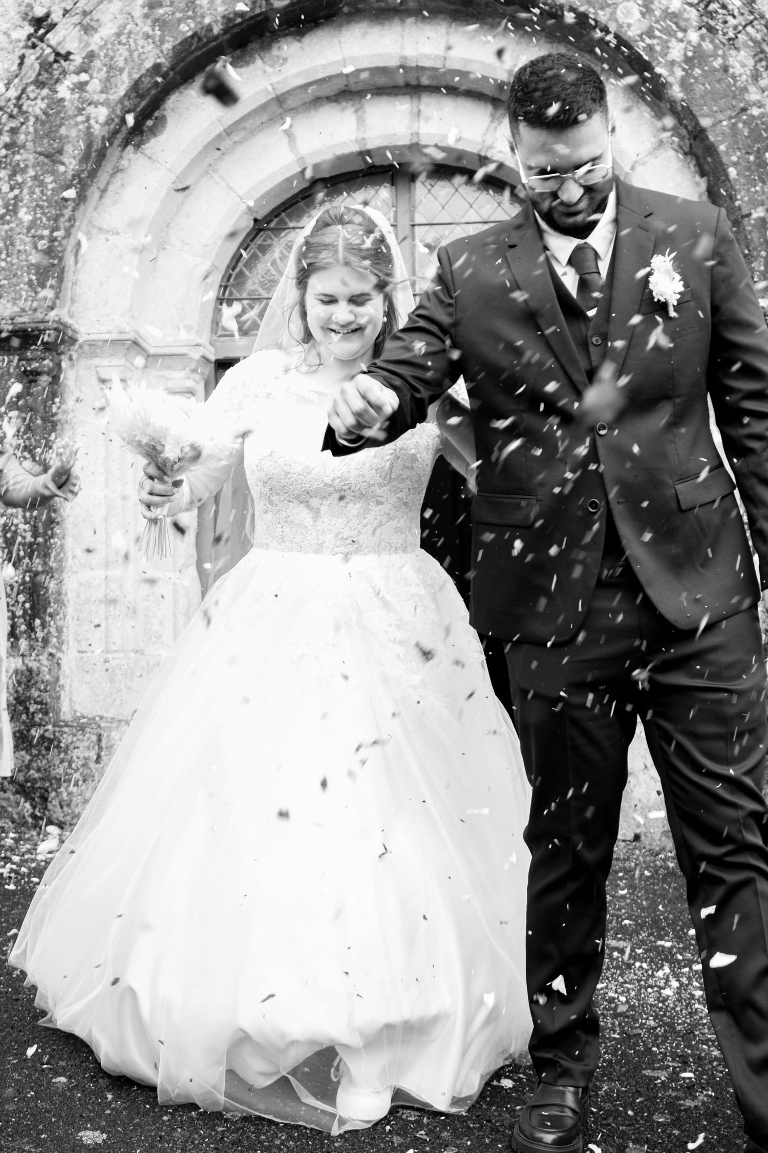 Black and white photo of a bride and groom walking through confetti outside a stone church after their wedding ceremony.