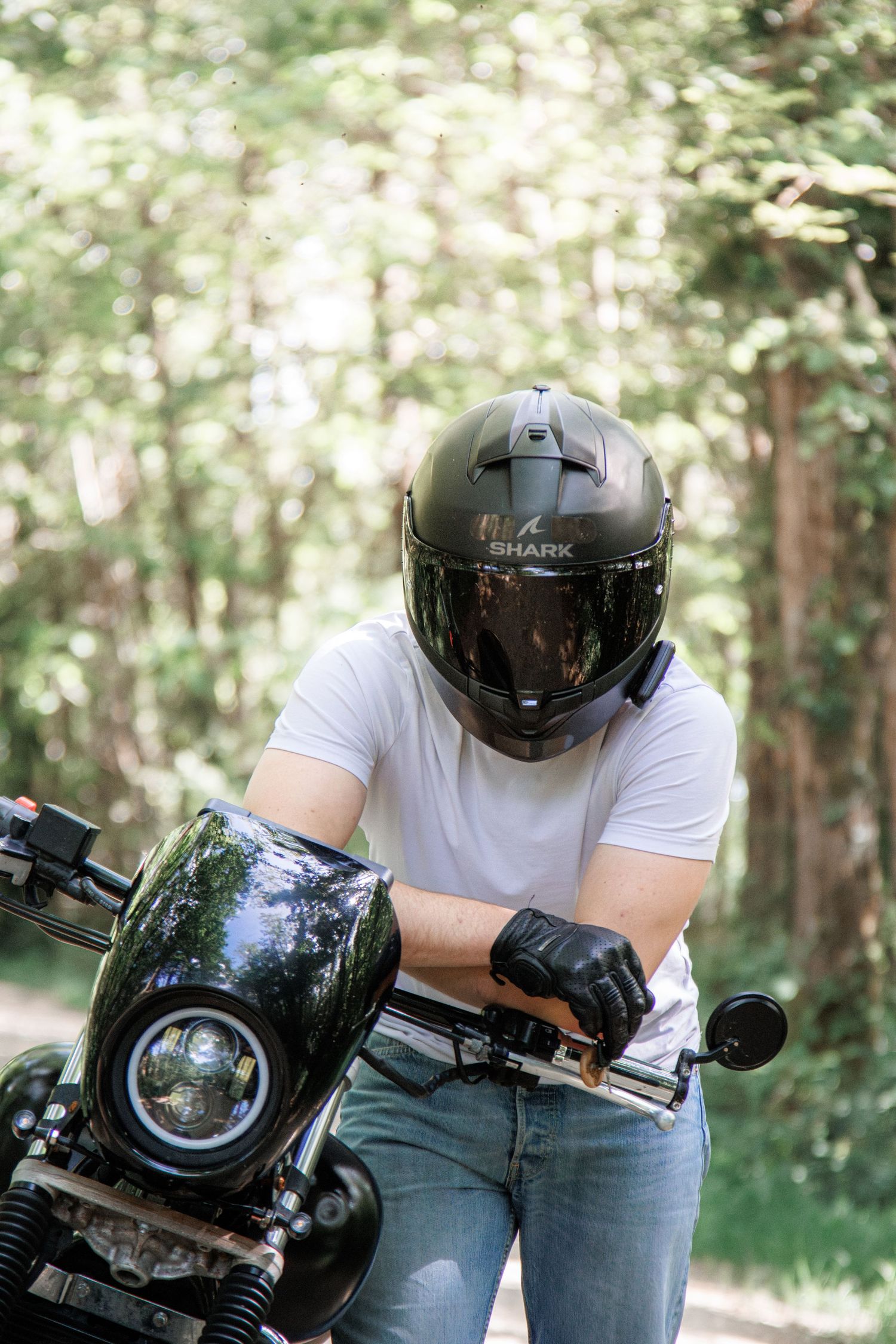Motorcyclist wearing a black Shark helmet and white t-shirt sitting on a dark green motorcycle in a forest setting.