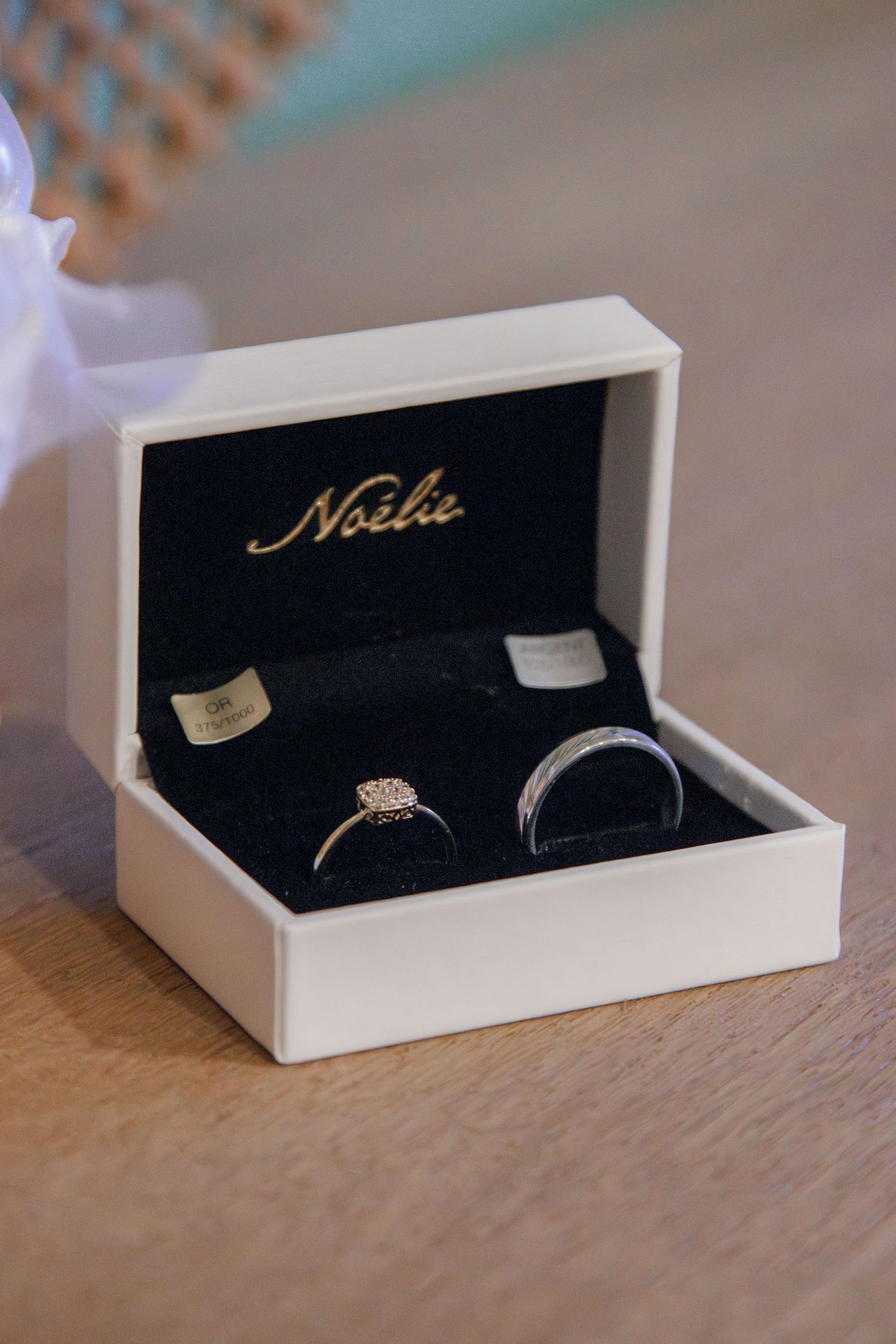 Two wedding rings displayed in an open white Noelie jewelry box with black velvet interior on a wooden surface.
