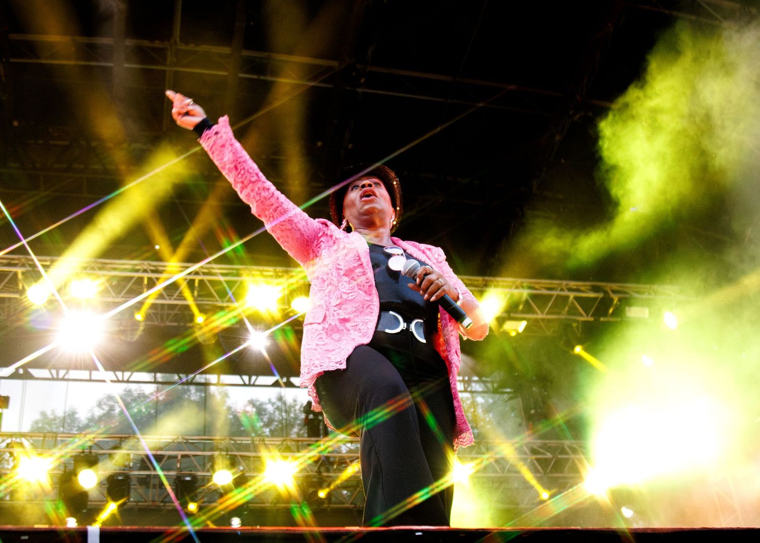 A performer on stage gestures dramatically under bright yellow stage lights and green smoke effects during a concert performance.