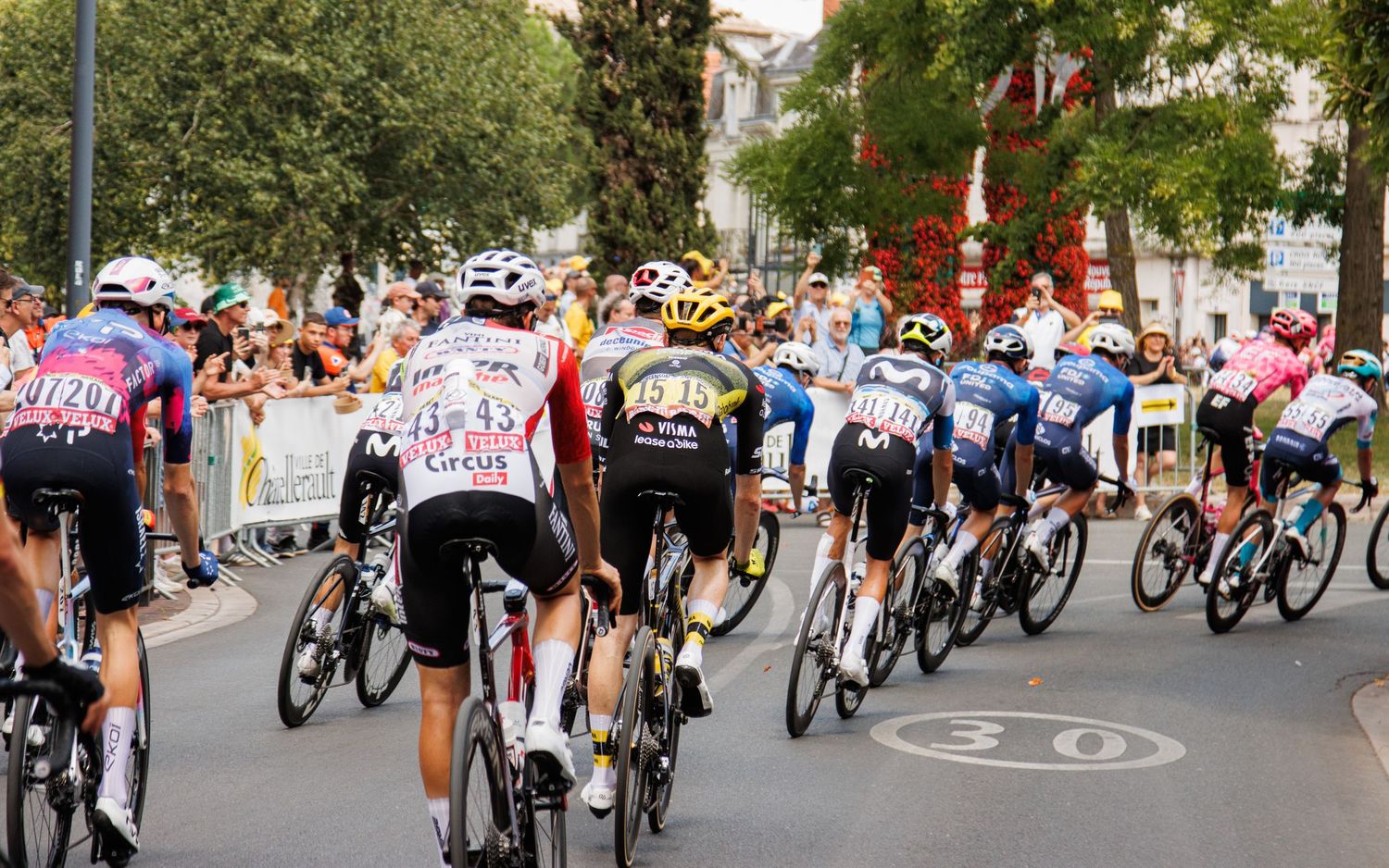 Professional cyclists race through a city street lined with cheering spectators during a cycling competition.