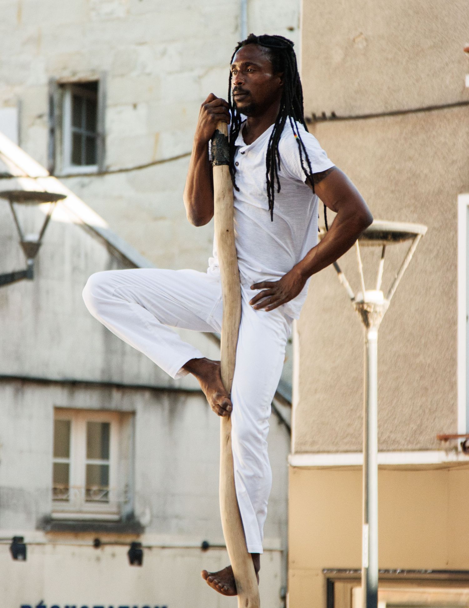 Performer in white clothing demonstrates balance skills while climbing a wooden pole during street show.