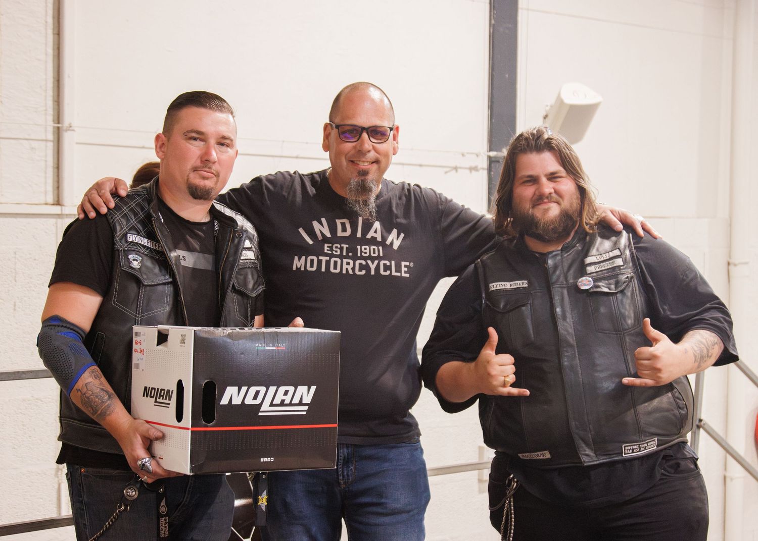 Three men pose with Indian Motorcycle branded merchandise in a warehouse setting.