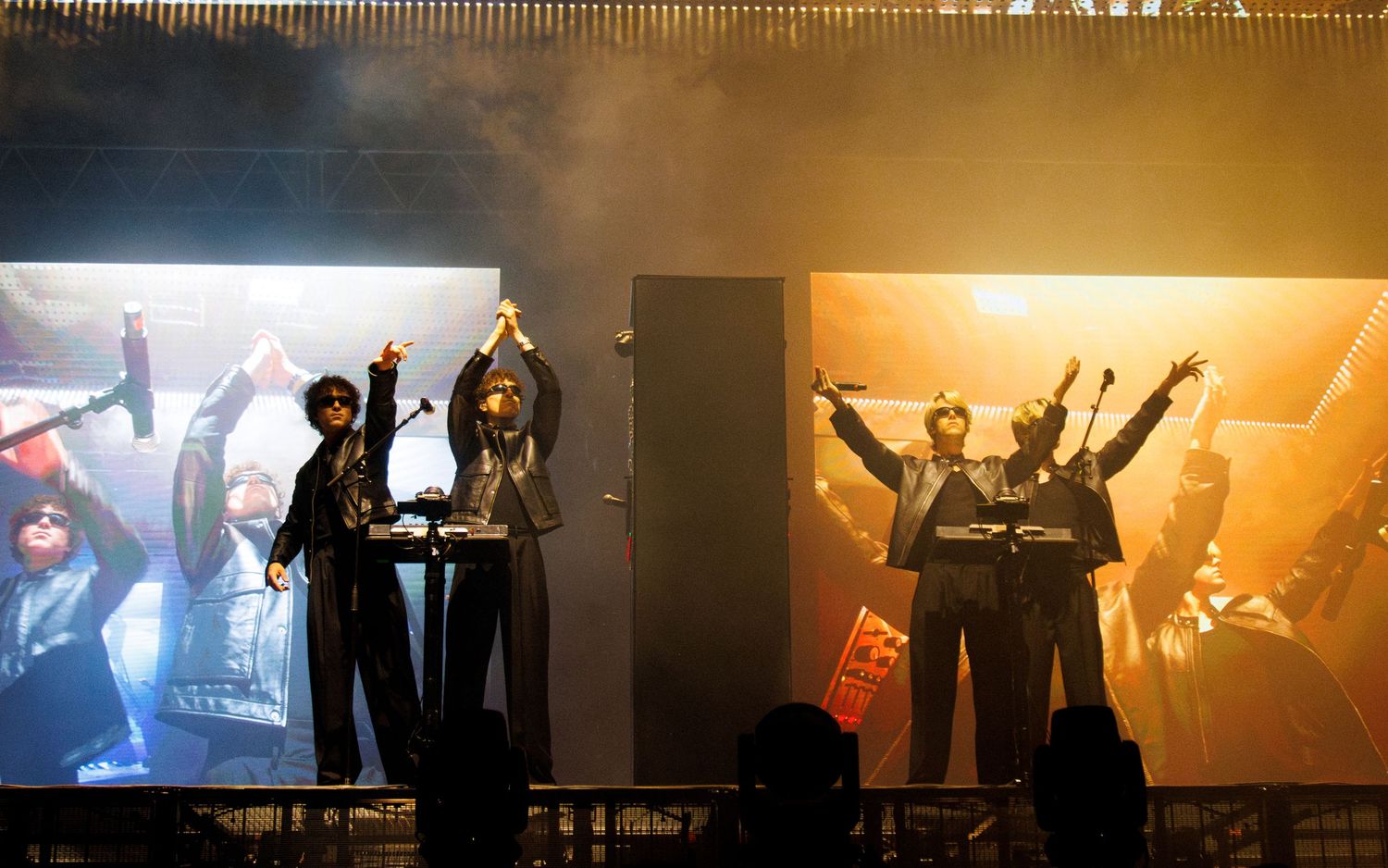 Silhouetted performers on stage raise their arms in celebration during a dramatic concert performance with orange lighting.