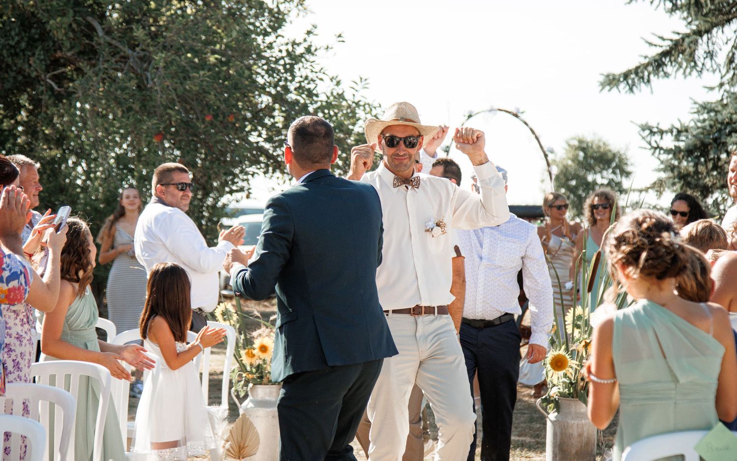 Wedding guests gather outdoors under trees for a summer celebration with people dancing and mingling.