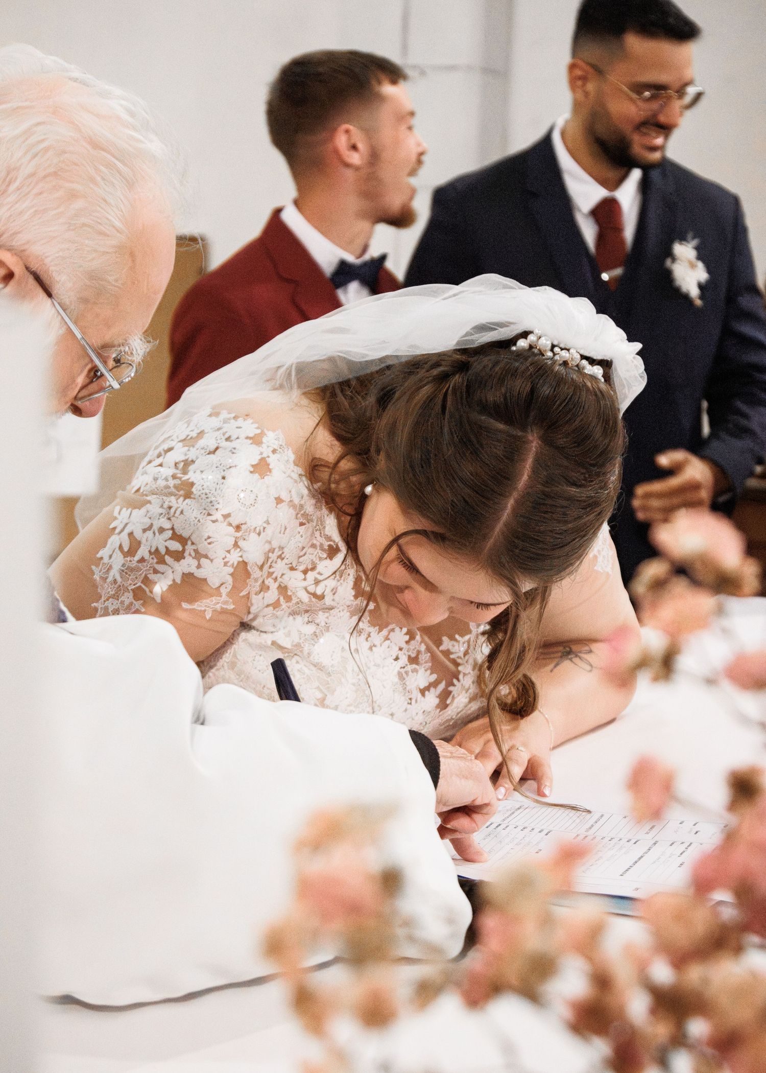 Bride in lace wedding dress and veil signing marriage certificate at ceremony table with guests in background.