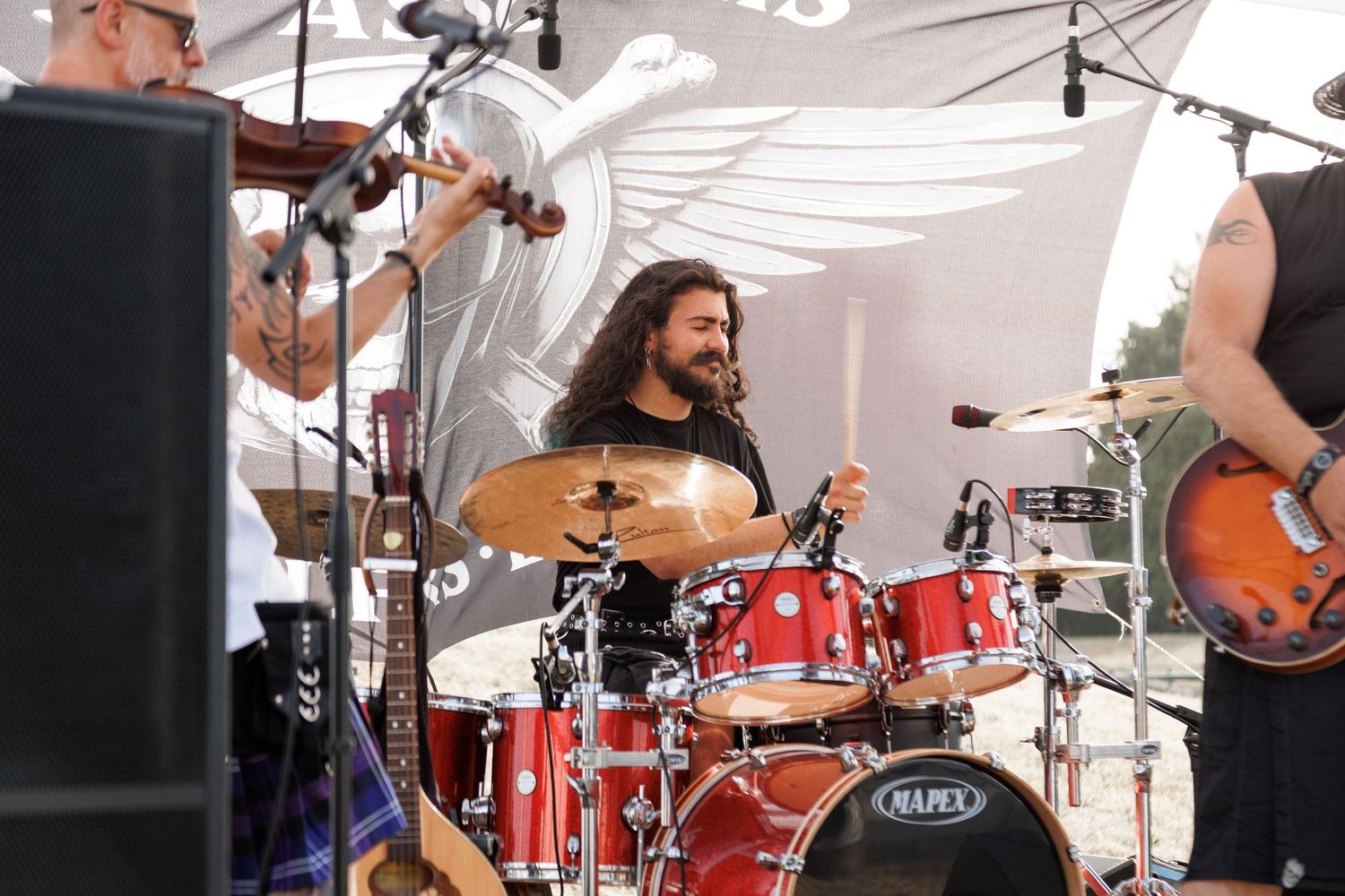 Smiling drummer plays red Mapex drum kit on outdoor stage with band members during live music festival performance.