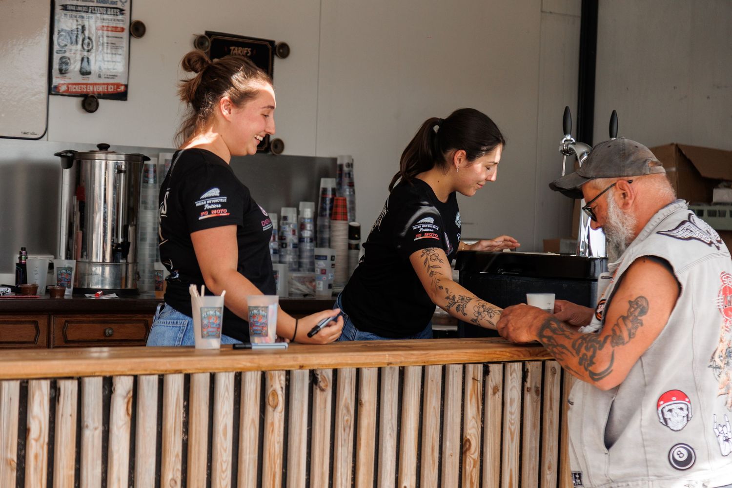 Two female baristas in black uniforms serve a tattooed male customer at a wooden bar counter.