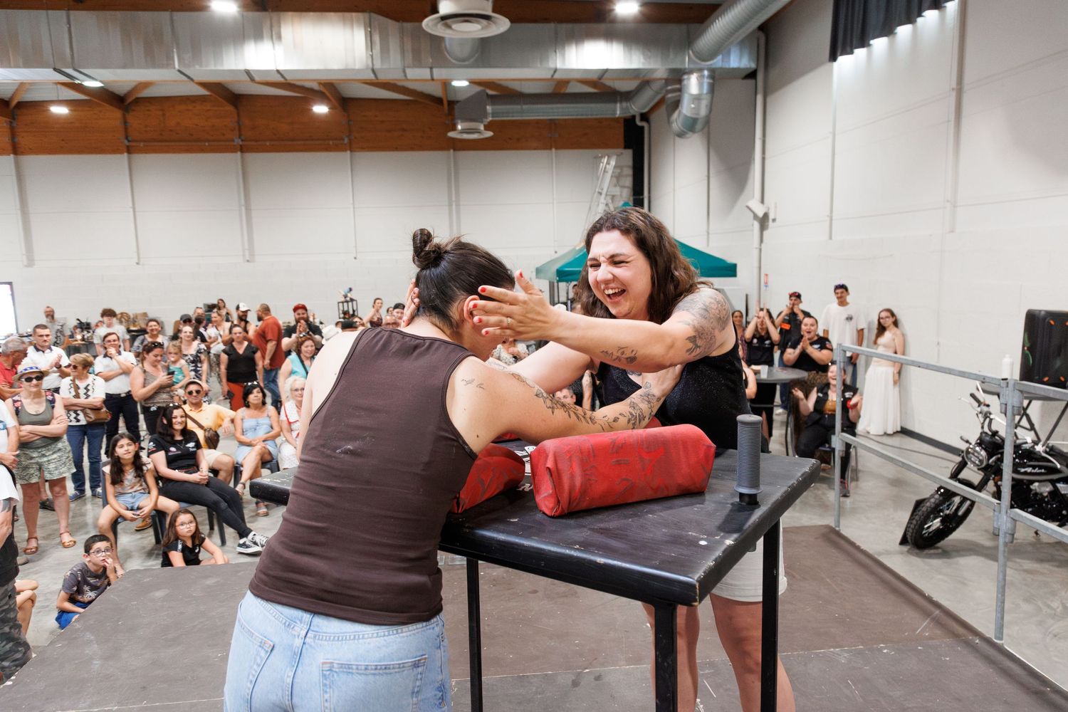 Two women arm wrestling on a padded table at an indoor event with a crowd of spectators watching.
