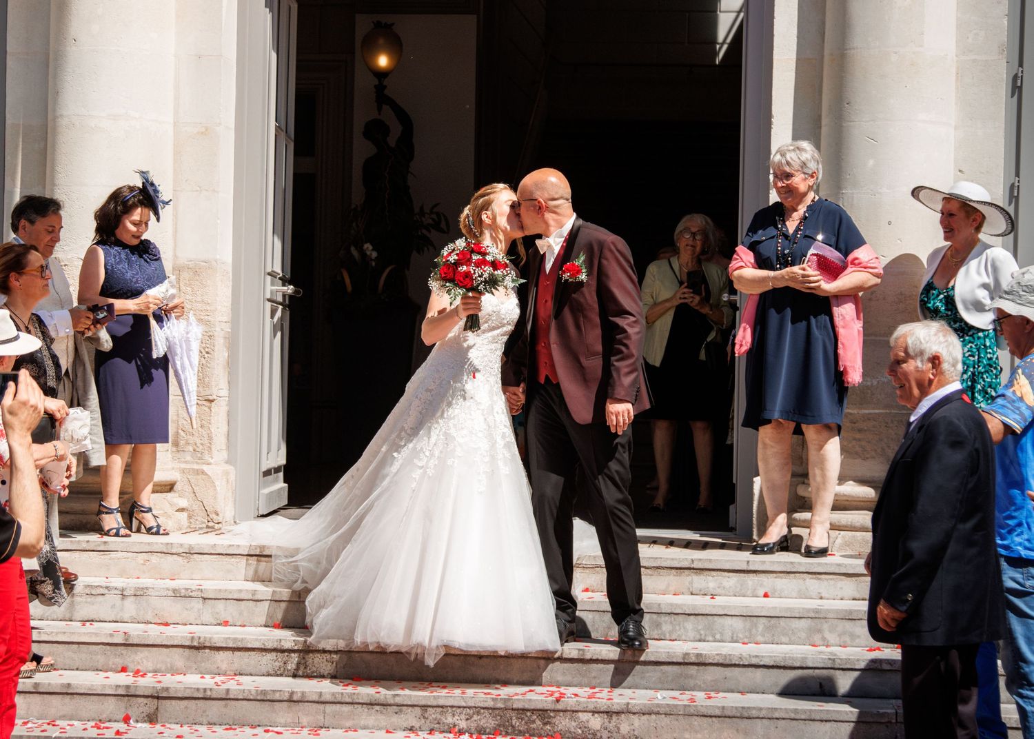A bride and groom share a kiss on the steps outside a venue as guests celebrate around them.